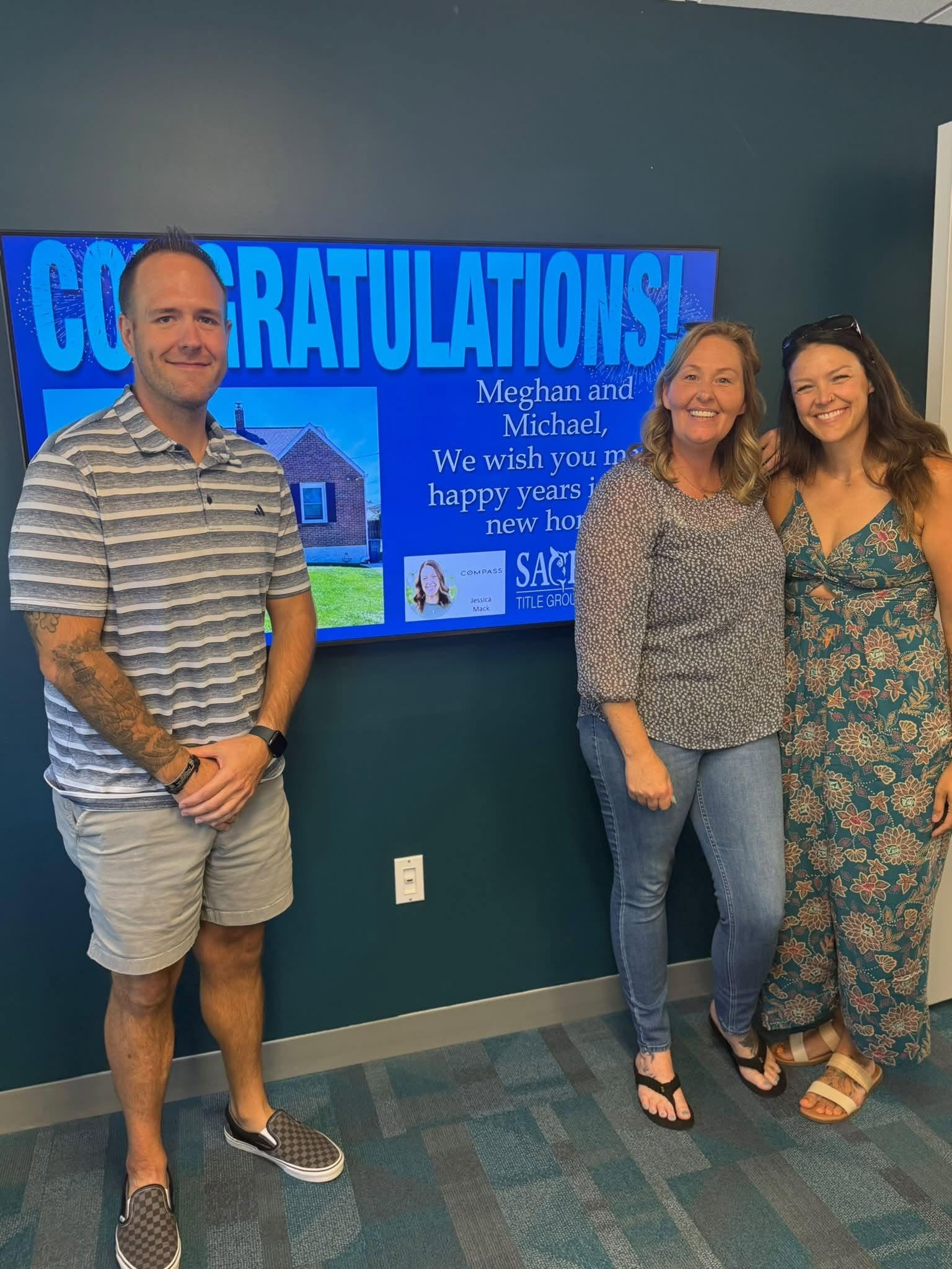 Three adults standing in front of a blue wall with a large screen displaying a congratulatory message. The man on the left is wearing a striped polo shirt, beige shorts, and checkered slip-on shoes. The woman in the middle is wearing a patterned blouse and jeans, and the woman on the right is wearing a floral dress and sandals. All three are smiling.