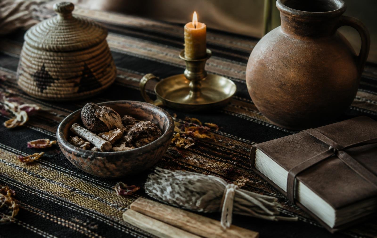 A rustic table setup with a bowl of dried herbs, a lit candle in a brass holder, a large brown ceramic jug, a wrapped journal or book, a small bundle of herbs, a wooden cutting board, and a teapot with a lid, all on a patterned woven cloth.