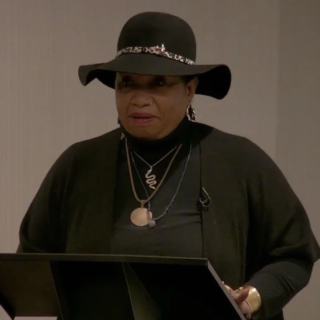 Mama Ayana wearing a wide-brimmed black hat with a ornament band, black top, and layered necklaces, standing beside a music stand.