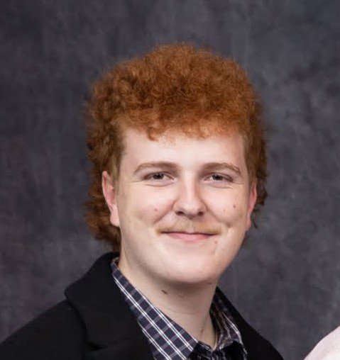 Portrait of a young man with curly red hair, smiling, wearing a dark blazer and a checkered shirt, against a dark textured background.