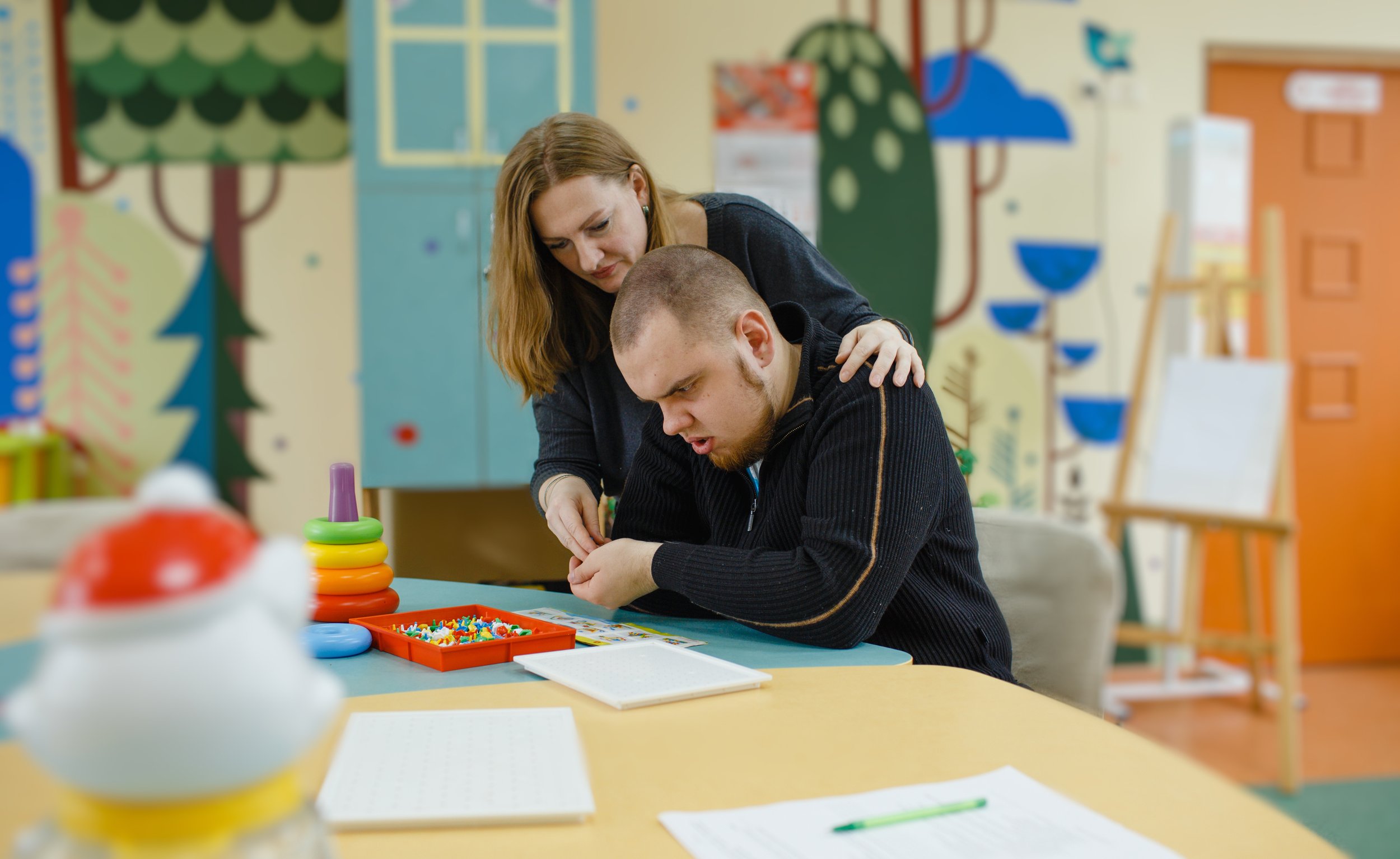 An adult helping a child with an activity at a colorful table in a classroom with playful wall decorations.