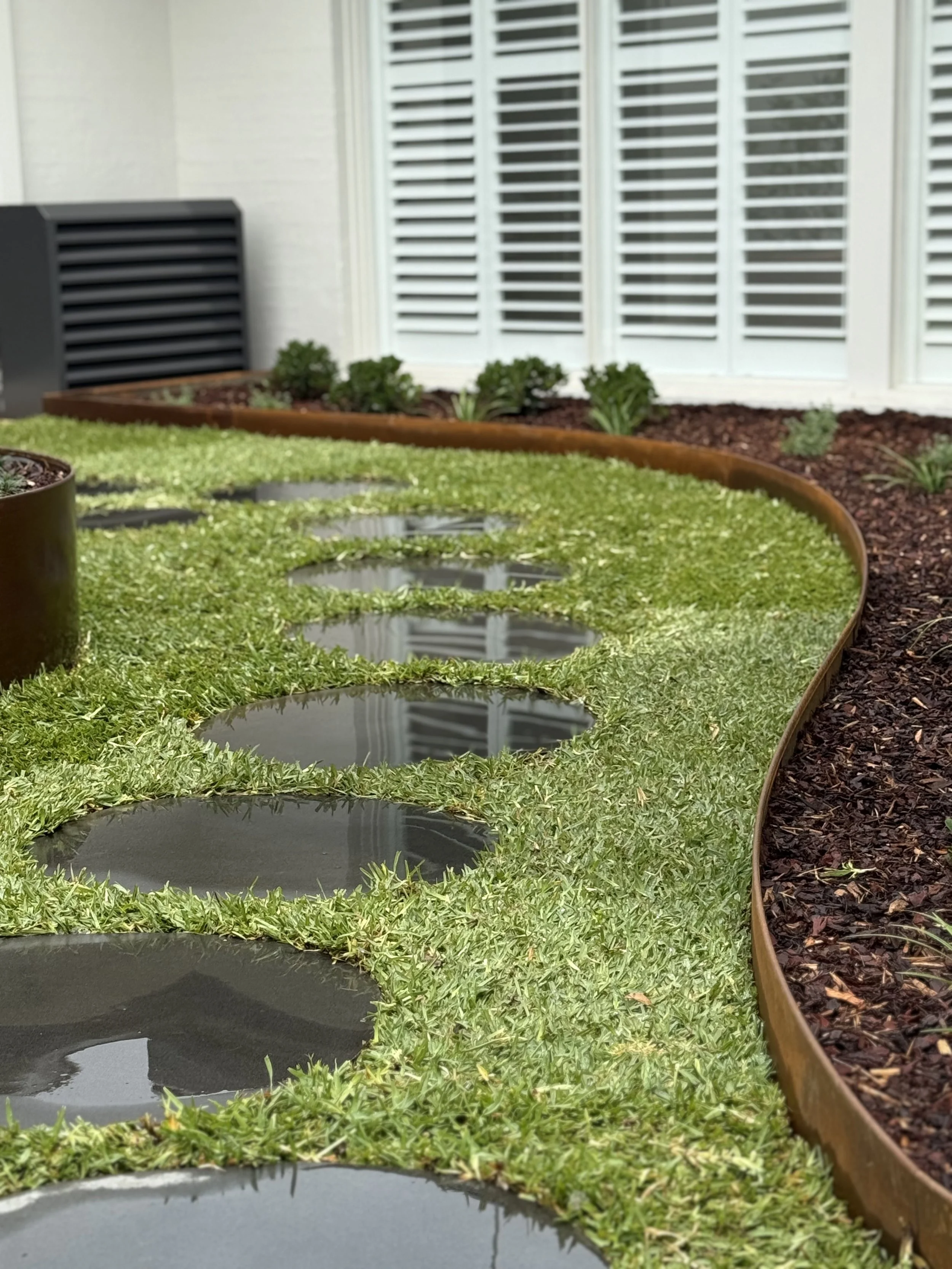 Close-up of a garden bed with water filled circular zones surrounded by green grass, with a white building with louvered shutters and a vent in the background.