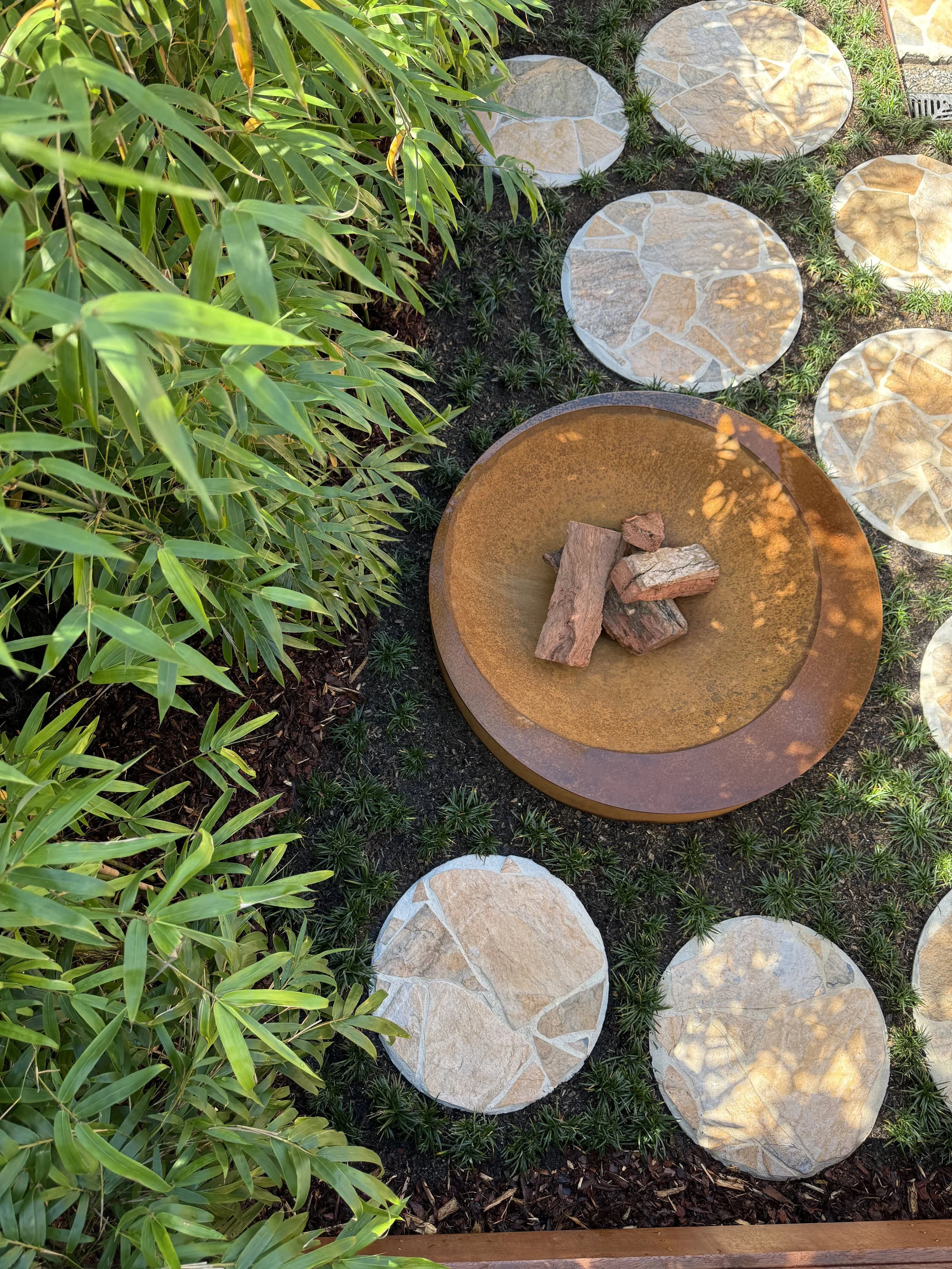 Garden pathway with large flat stones surrounded by small green plants and a bush with long green leaves, with a circular rust-colored fire pit containing rocks in the center.
