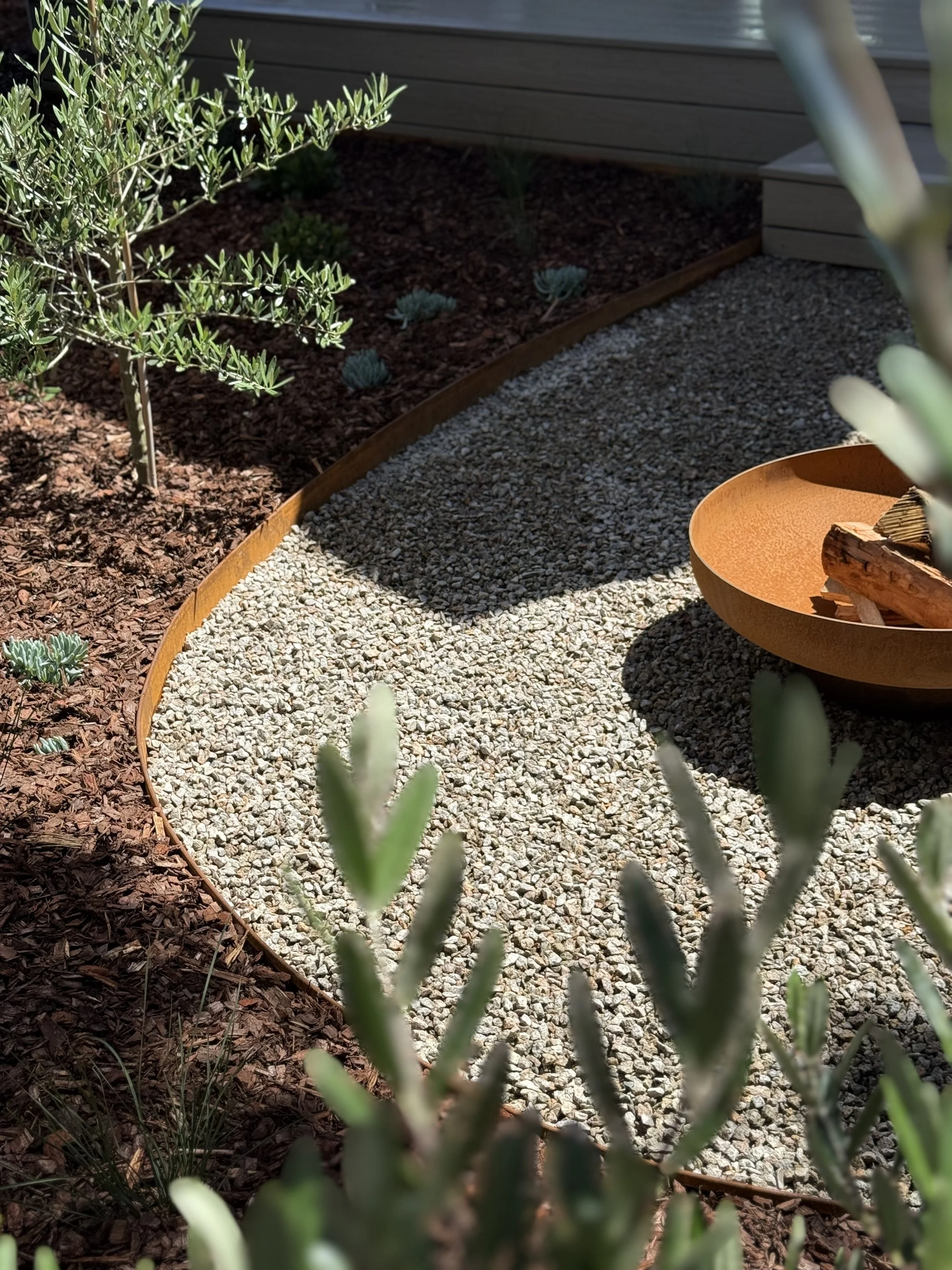 A garden with a variety of plants, a gravel path, and a decorative shallow bowl with wooden pieces inside.