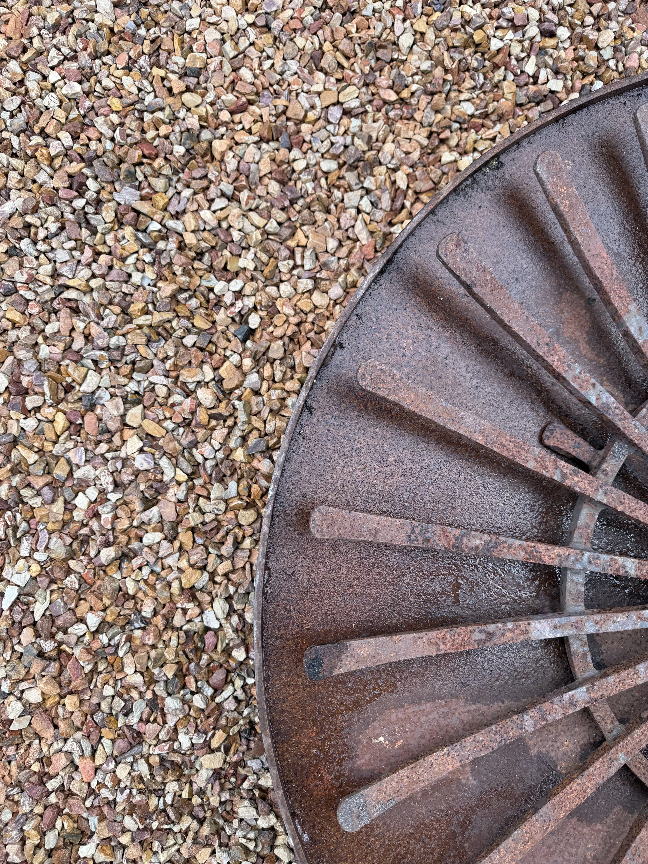 Close-up of a rusted metal grill or grate with evenly spaced slats, situated on a bed of small, multi-colored gravel stones.
