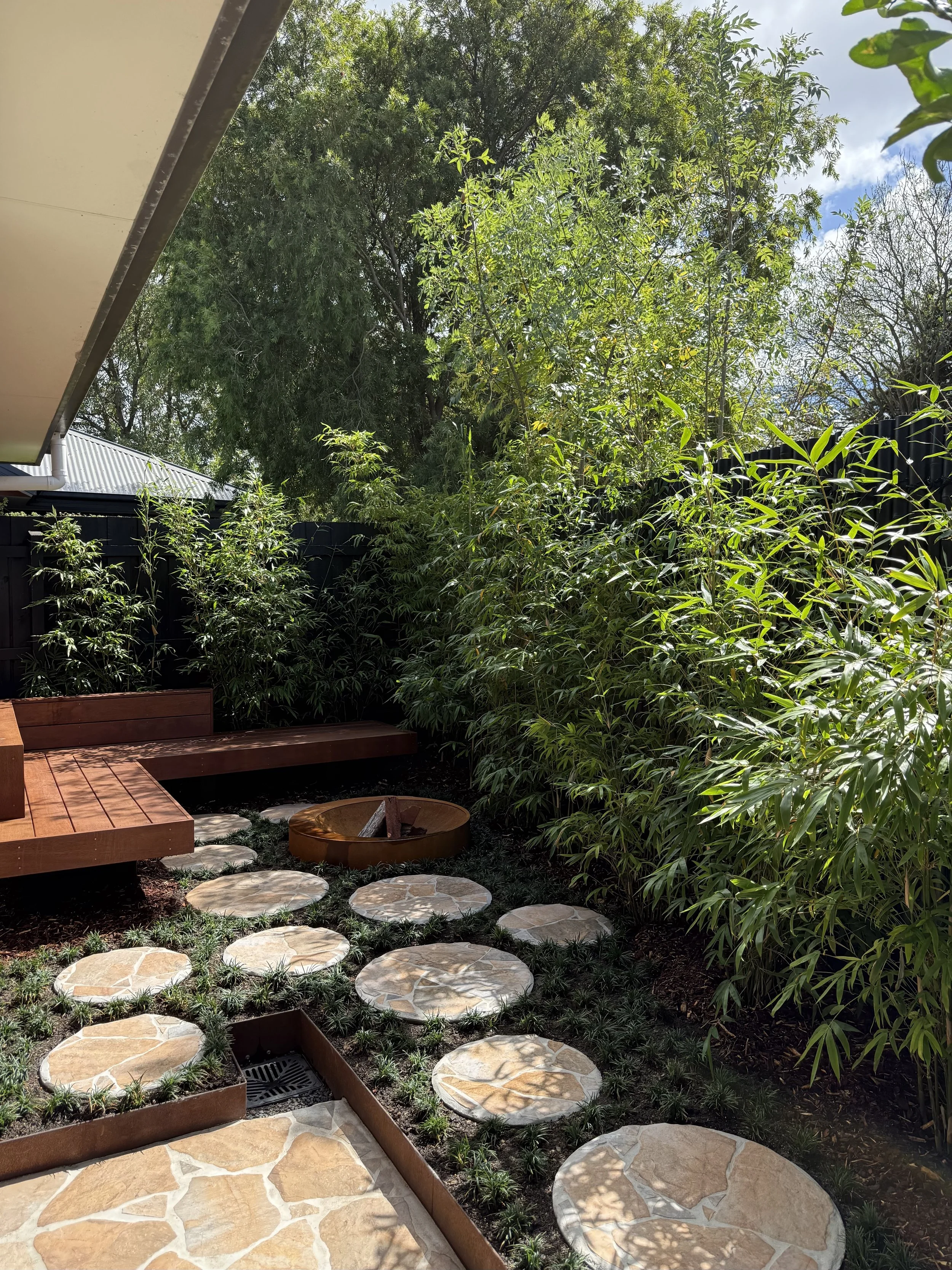A backyard garden with various green plants and trees, a wooden bench, circular stepping stones on the ground, and a fire pit. Part of a house roof and gutter are visible in the top left corner.