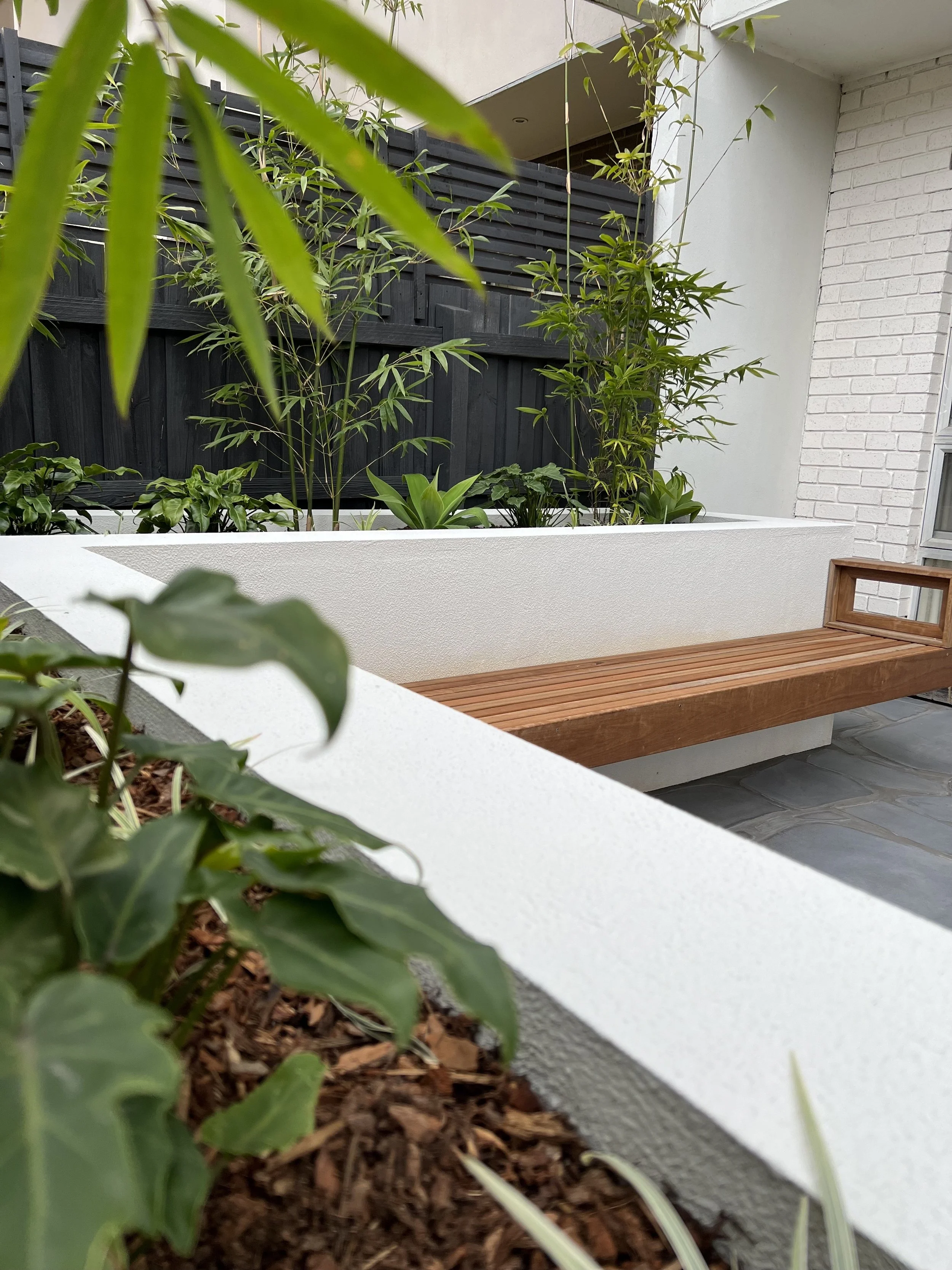 Close-up of a modern outdoor patio garden with a white concrete planter containing green foliage, including leafy plants and a tall bamboo, and a wooden bench on a stone-tile floor against a white wall with a dark wooden fence in the background.