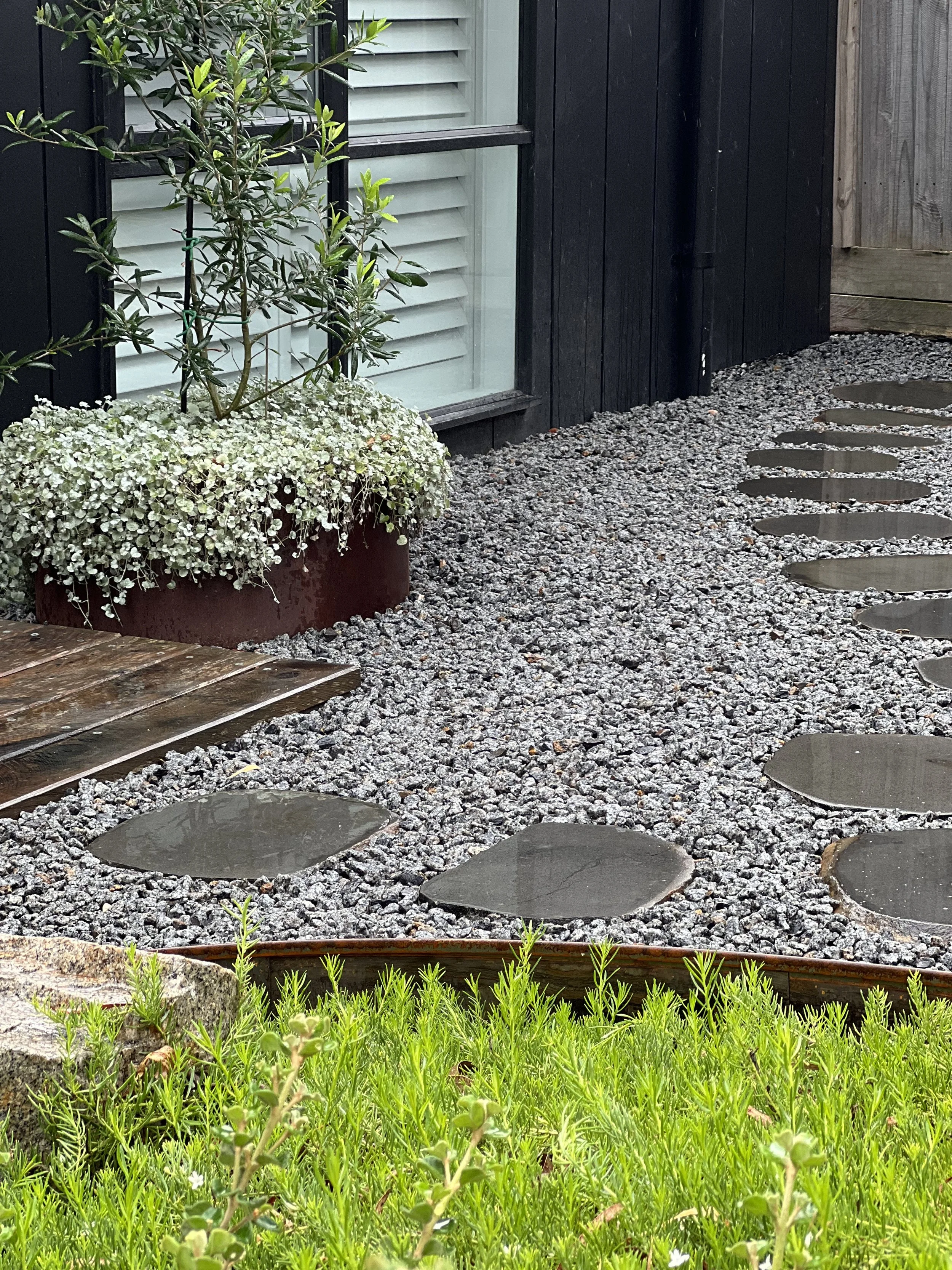 A small garden with green grass and small flowering plants in the foreground, a gravel pathway with round stepping stones on the right, and a black wooden fence with a window, a plant in a large pot, and a wooden fence on the background.