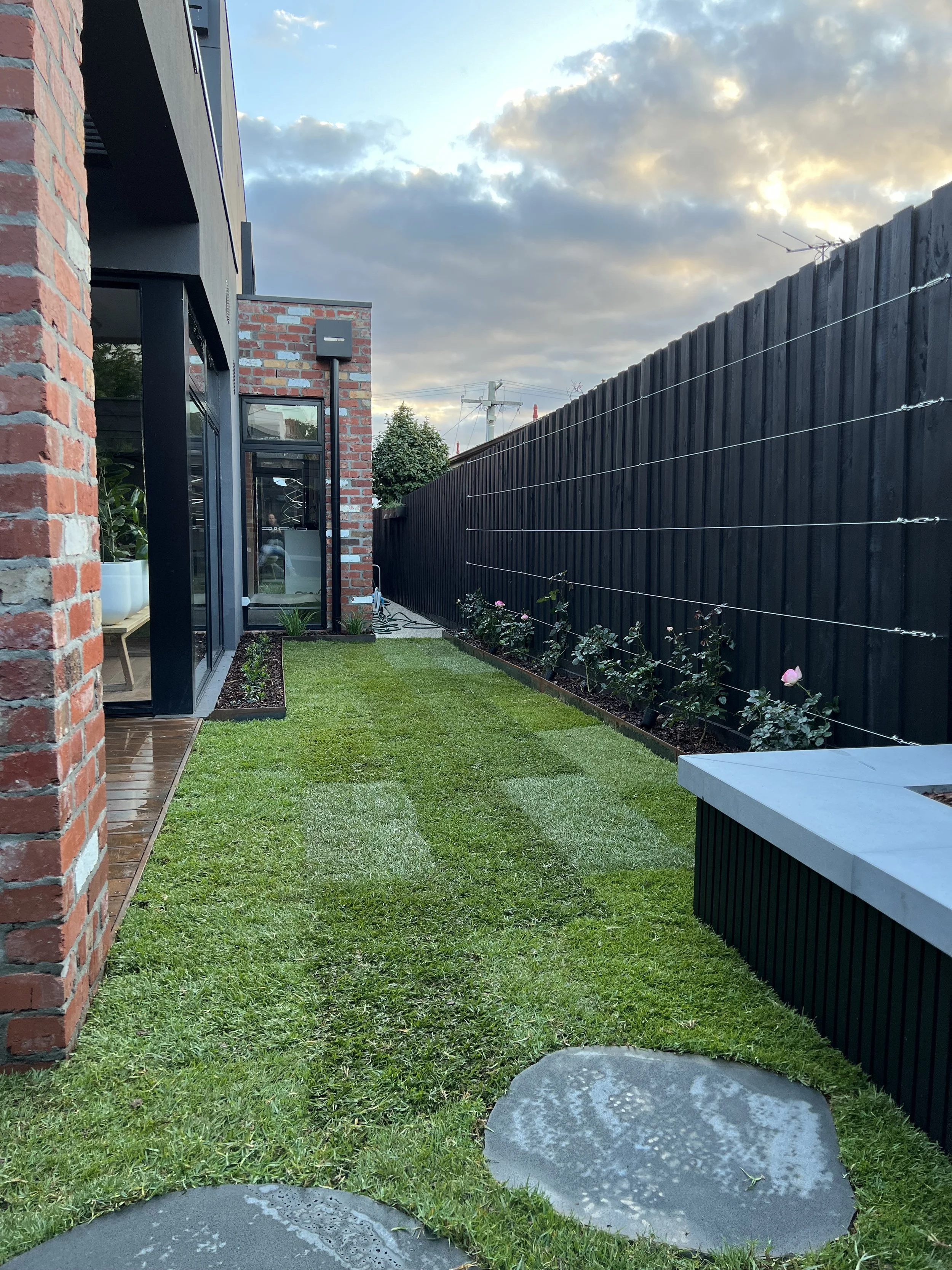 A backyard with a small grassy area, a brick house on the left, a black wooden fence on the right, and a cloudy sky above. There are a few plants along the fence and some round stepping stones on the grass.