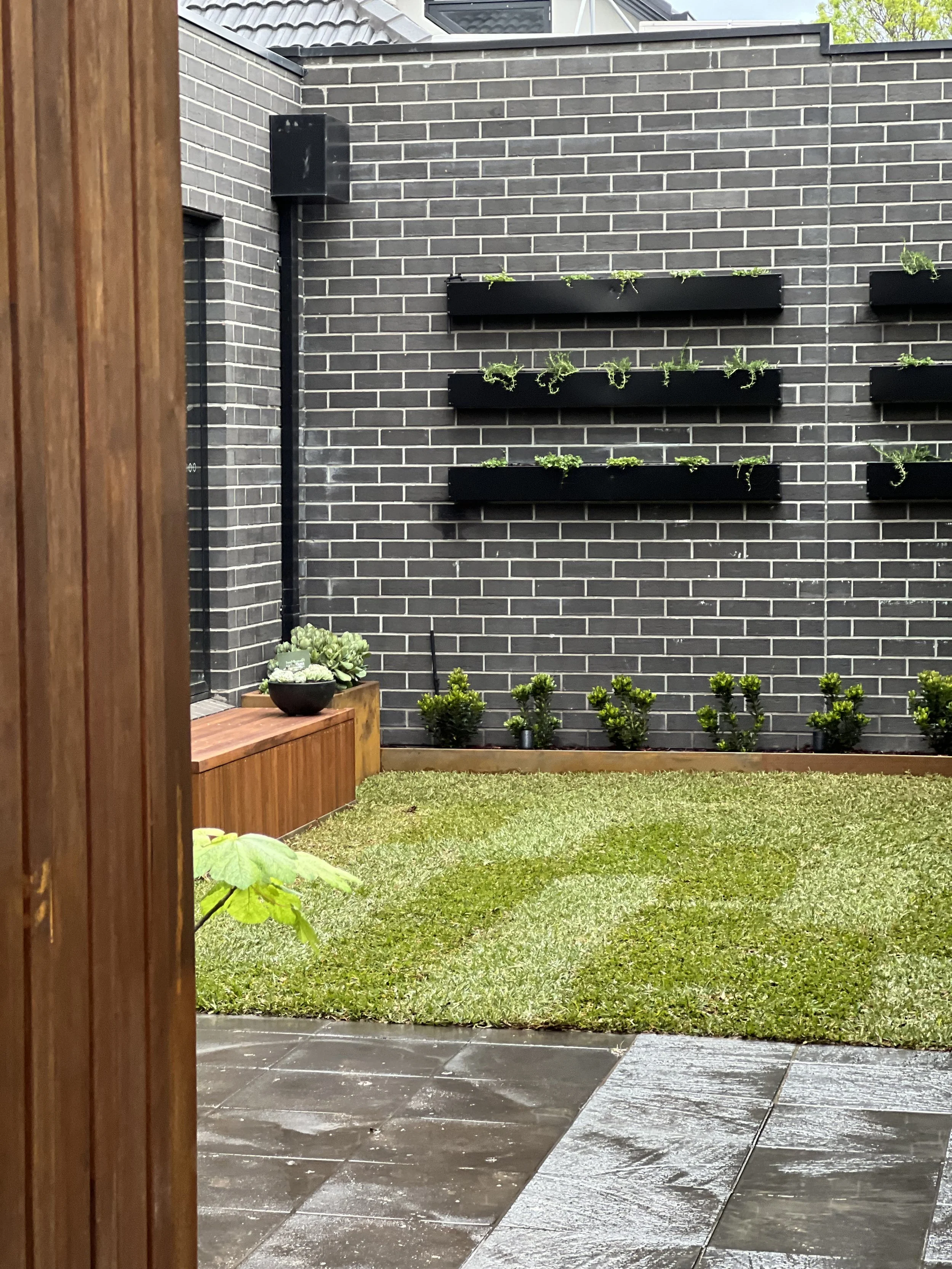 View of a backyard garden with a freshly mowed grass lawn, black brick wall, horizontal black planters with green plants, a wooden bench with potted plants, and a wet stone patio.