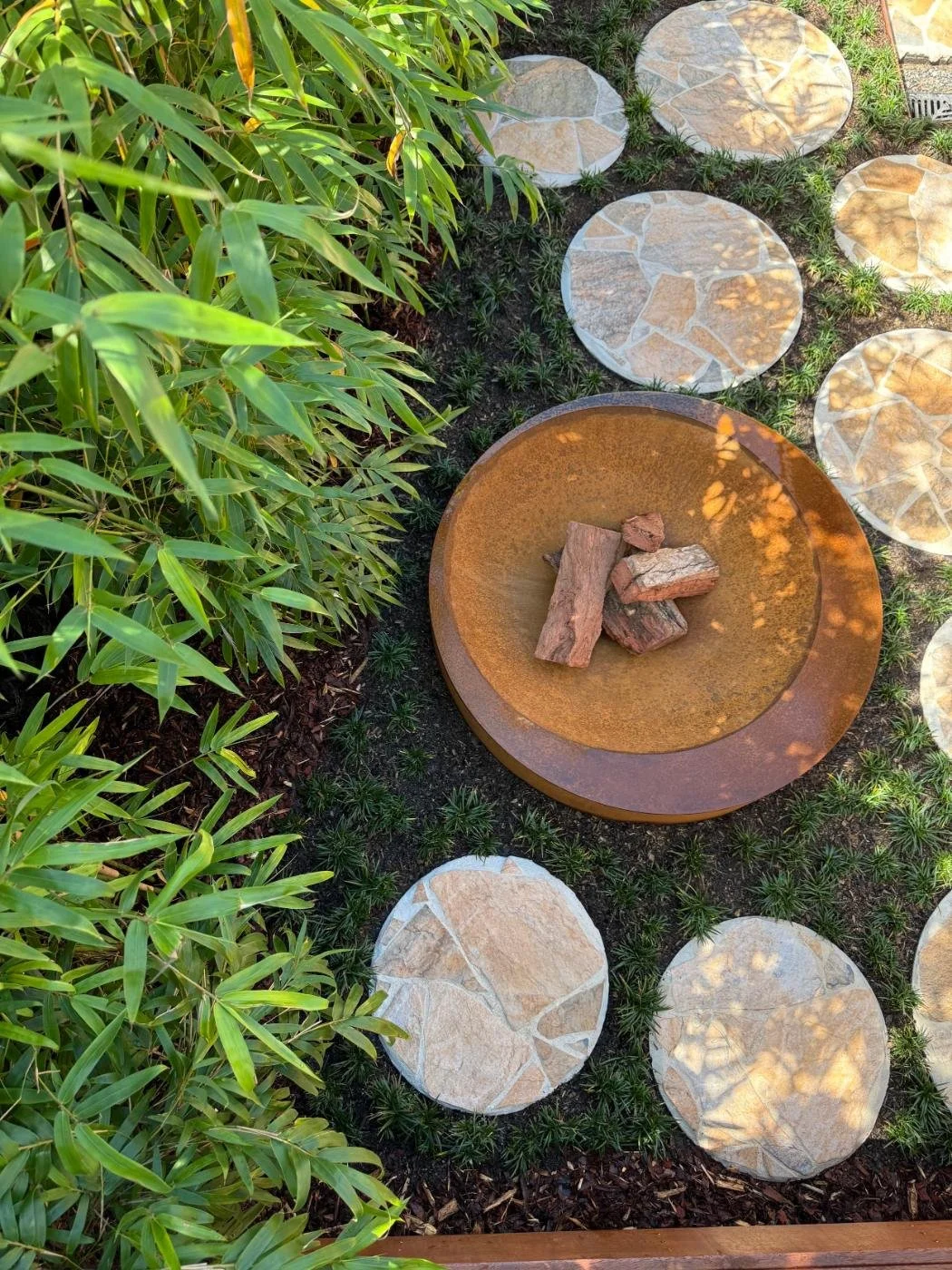 Garden pathway with large, flat, circular stone stepping stones embedded in dark soil. To the left, green leafy bushes. In the center, a rust-colored fire pit with rocks inside. In the background, small green plants and a drain grate visible.