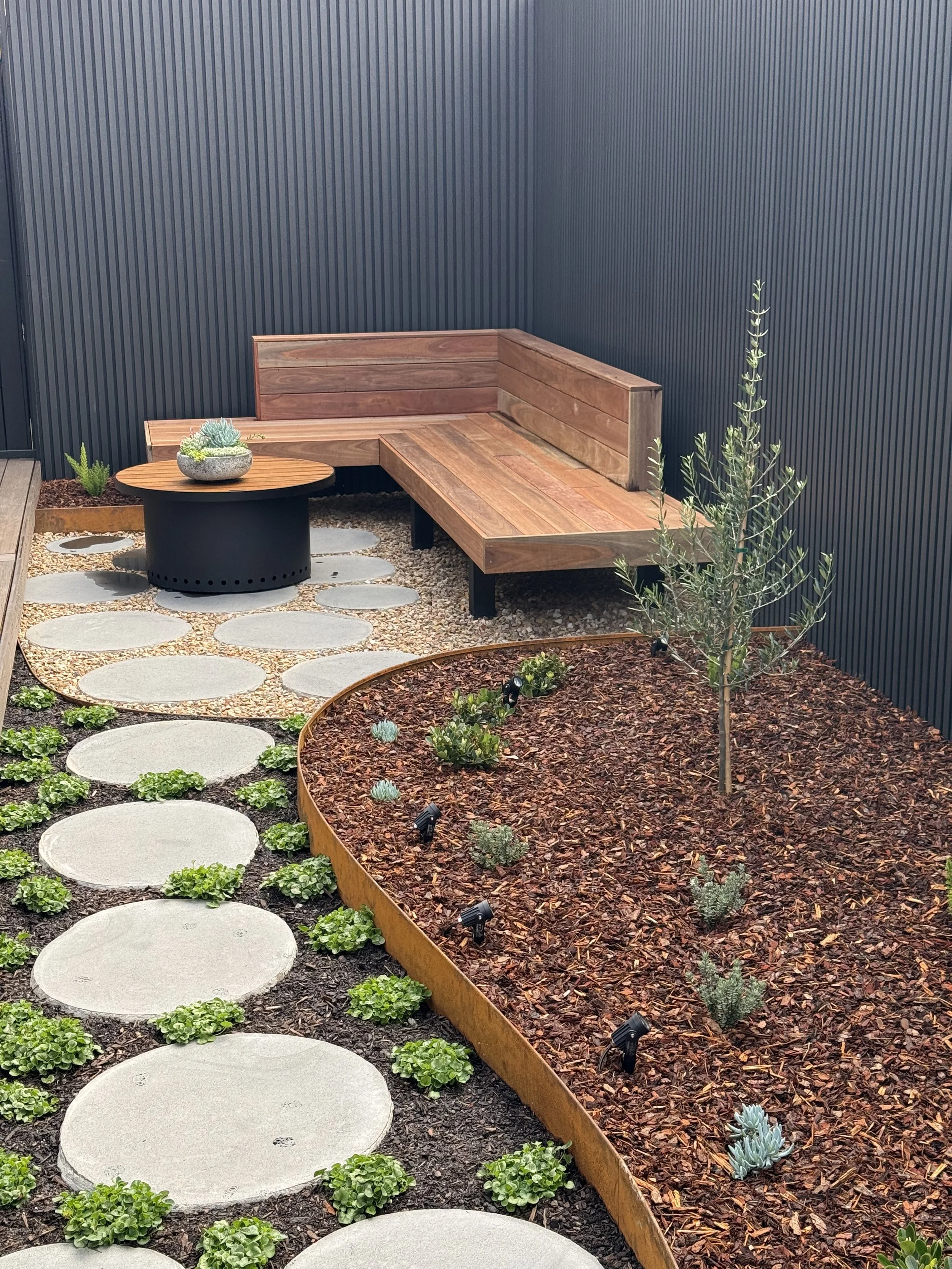 A small outdoor garden area featuring a wooden corner bench, a round black fire pit table with a succulent arrangement, a pathway of large circular stepping stones with greenery, and a raised flower bed with young plants and mulch, enclosed by dark-c