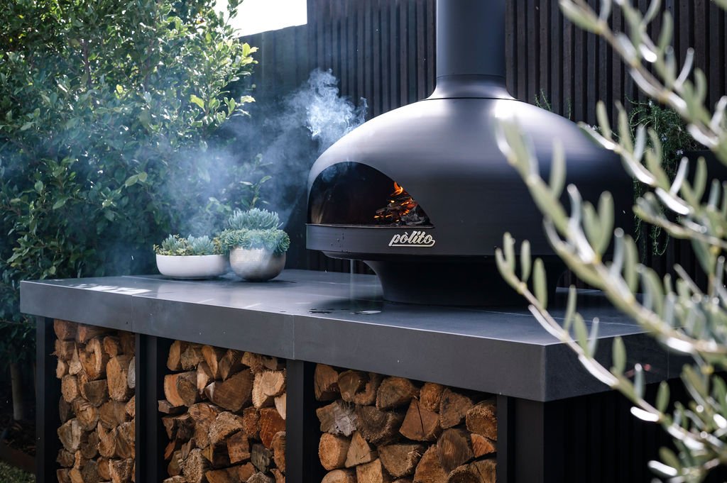 A black pizza oven with the brand name Politto, placed on a gray countertop, with small fire glowing inside, emitting smoke, and surrounded by green foliage and a stack of firewood underneath.