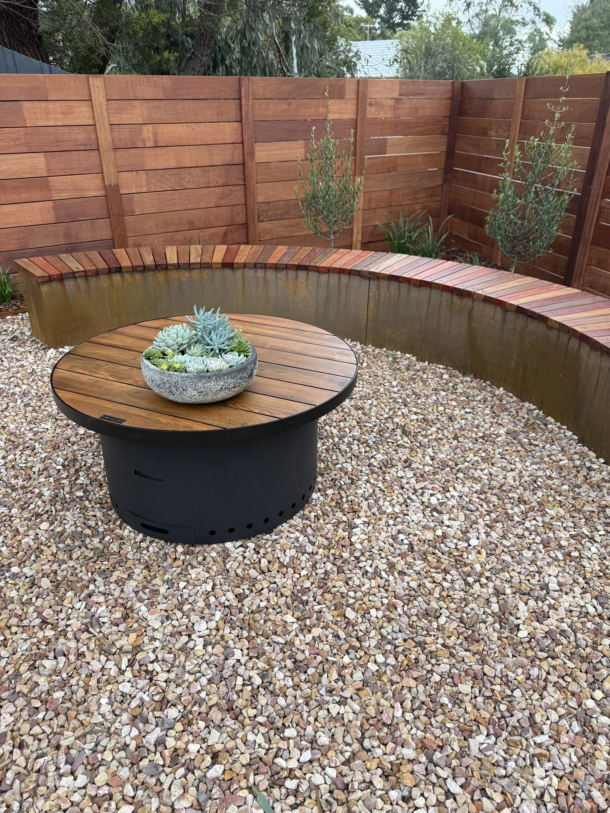 A backyard patio with a rotating fire pit table on gravel ground, a curved wooden bench, a tall plant, and a wooden privacy fence, with trees and house rooftops in the background.