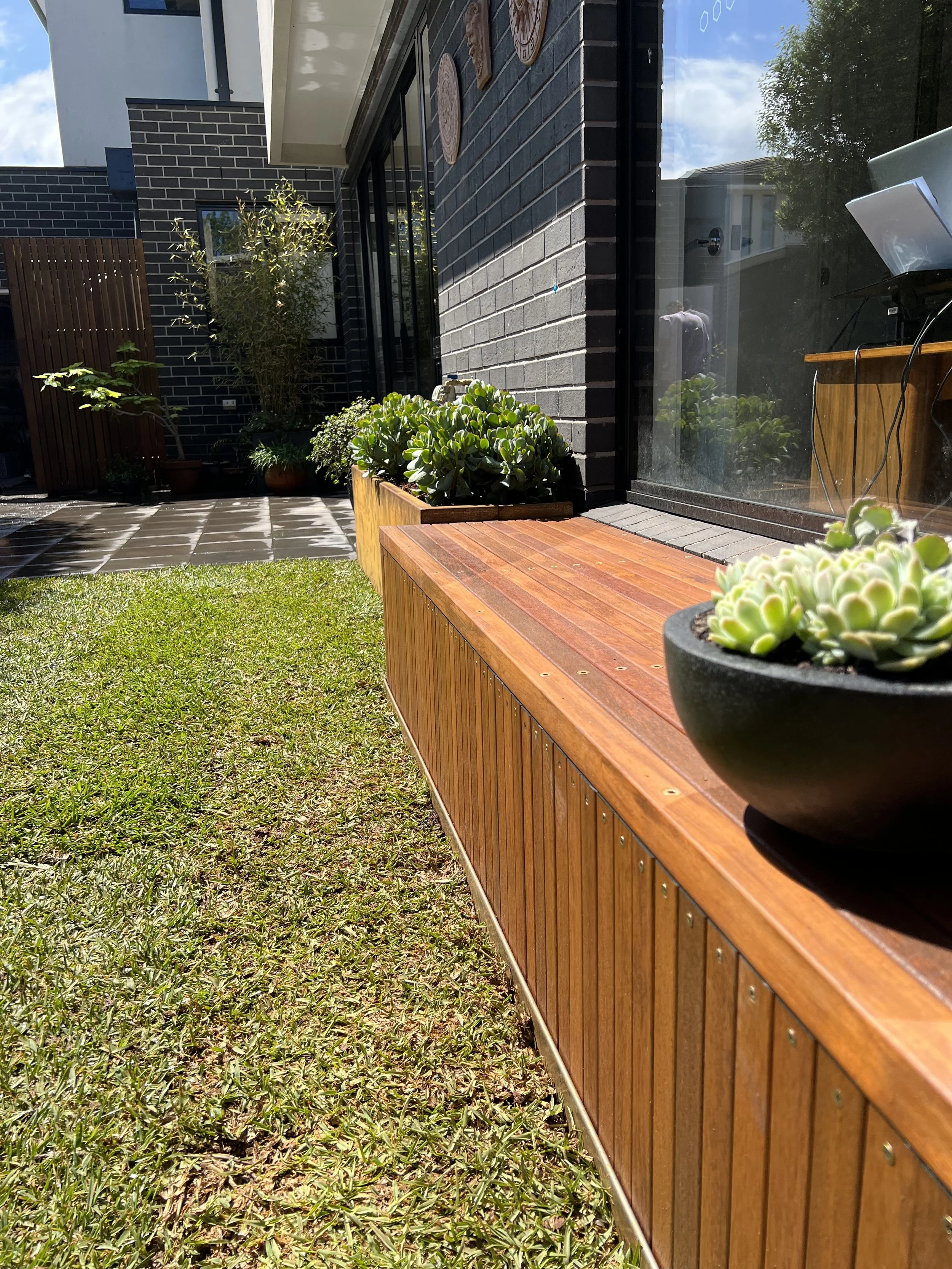 A backyard patio area with a large wooden planter filled with succulent plants, a grassy lawn, and a glass sliding door leading into a house. There are potted plants, a small tiled section, and some wall art outside.