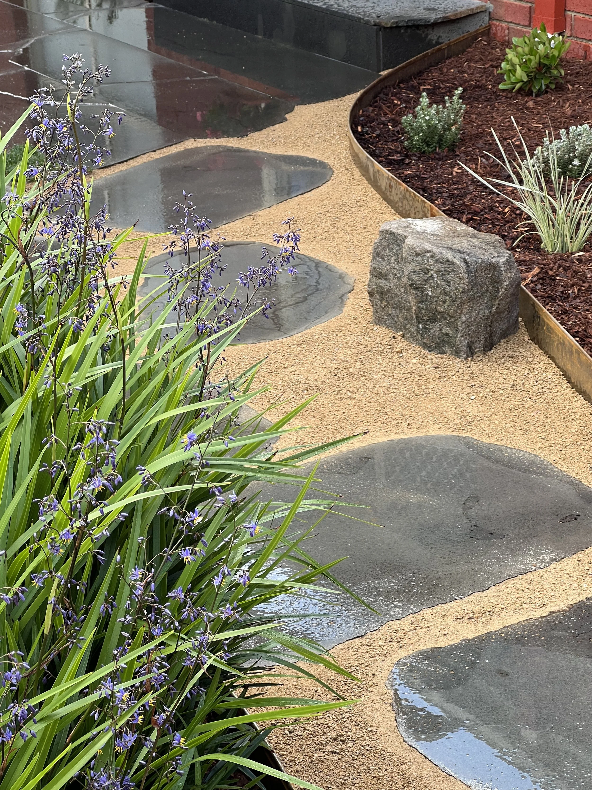 A garden pathway with flat stones, a small stream of water, green plants, and a rusted metal border around a flower bed with mulch and small green plants, with a brick wall in the background.