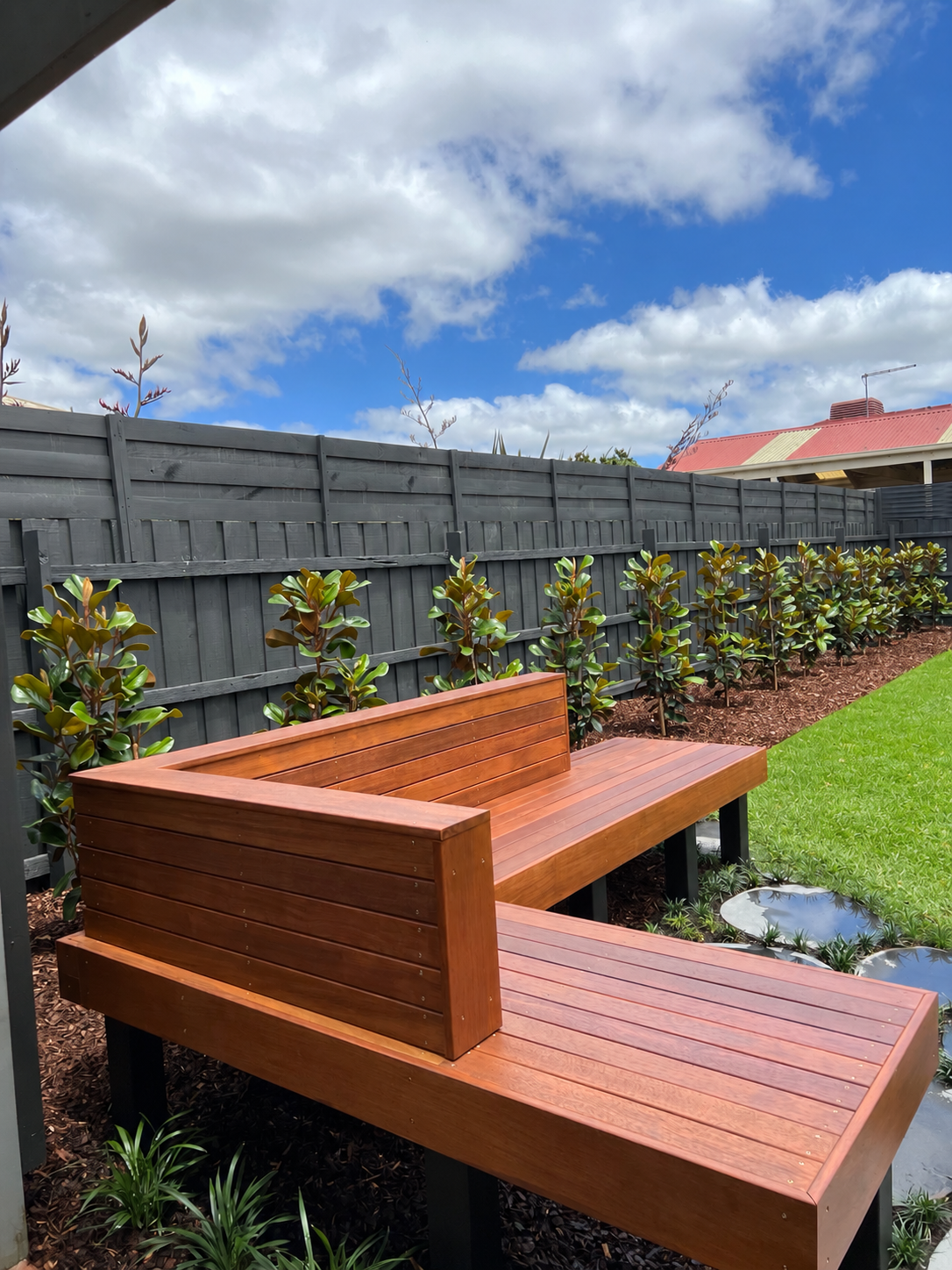 A backyard with a modern wooden bench, a row of bushy green plants, a dark gray wooden fence, and a partly cloudy sky.
