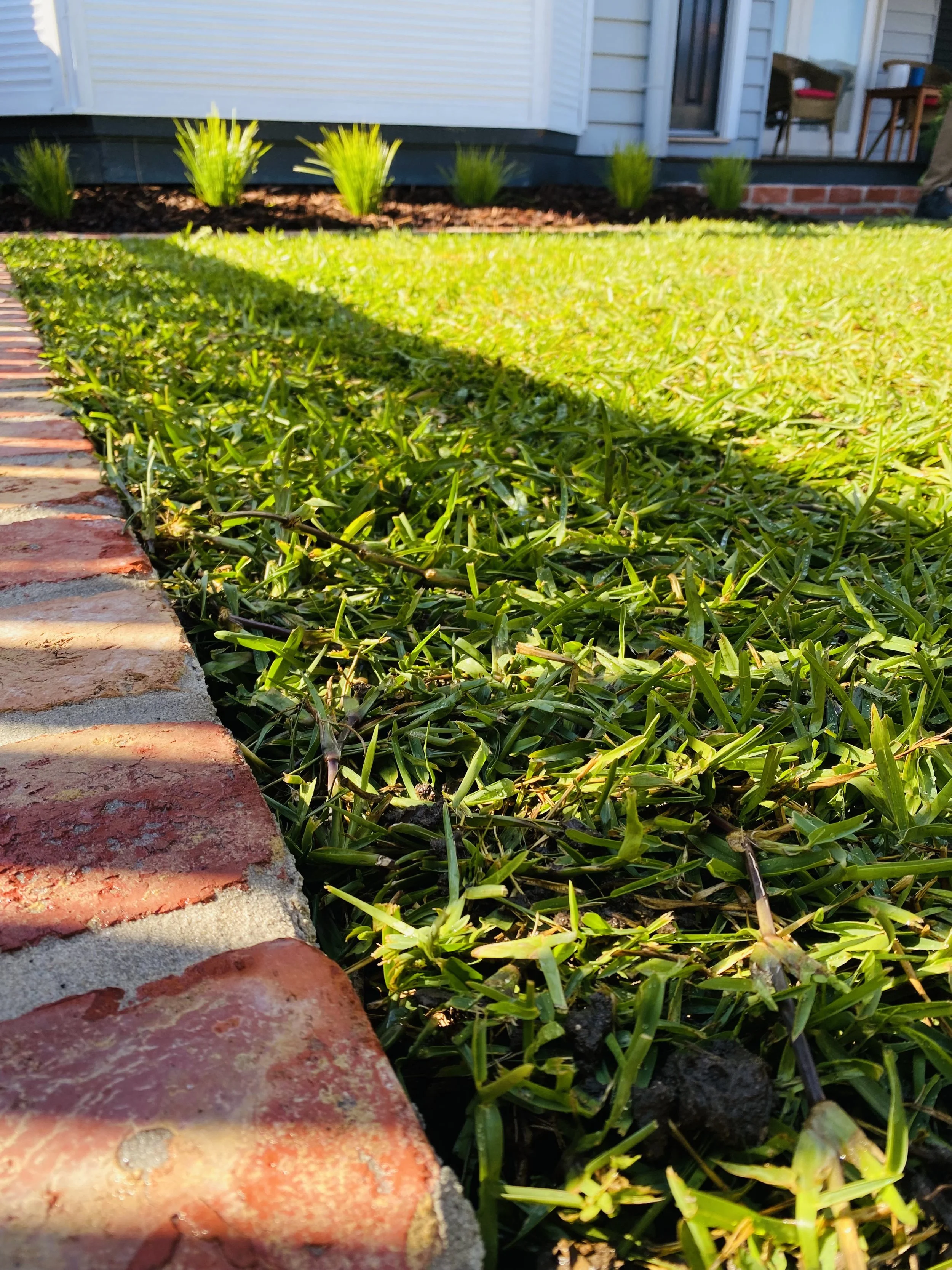 Close-up view of a brick pathway and a green lawn with small bushes, part of a house exterior with a porch and outdoor furniture in the background.