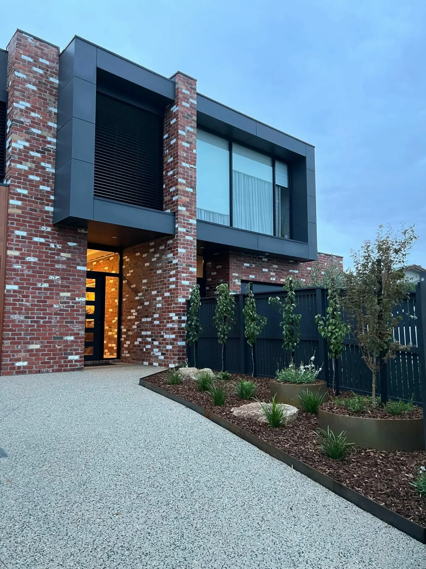 Modern house with red brick and black metal accents, front yard with small plants and trees, gravel driveway, overcast sky.