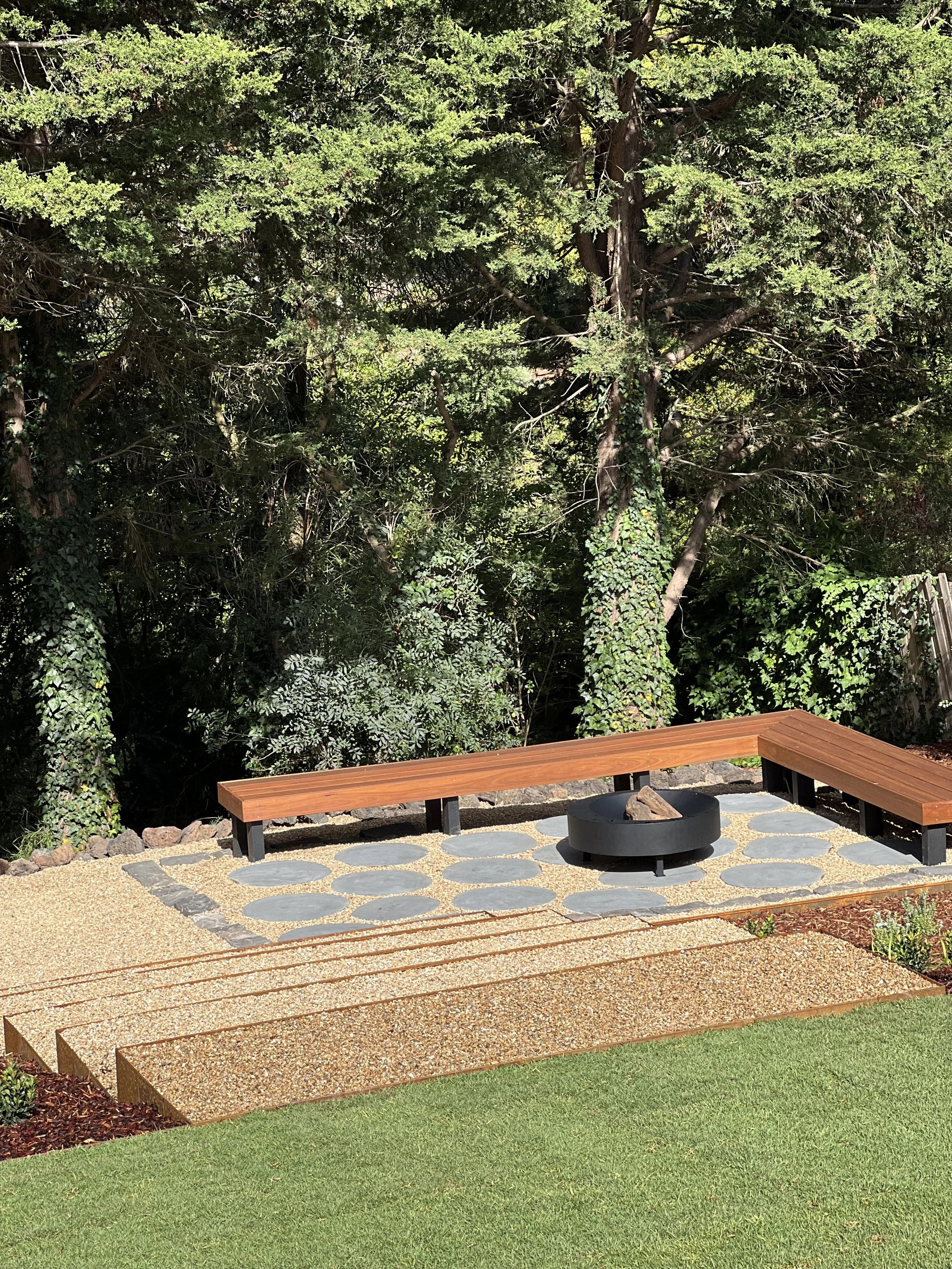 Outdoor seating area with a wooden bench, a fire pit, and a gravel surface, surrounded by trees and a grassy lawn.