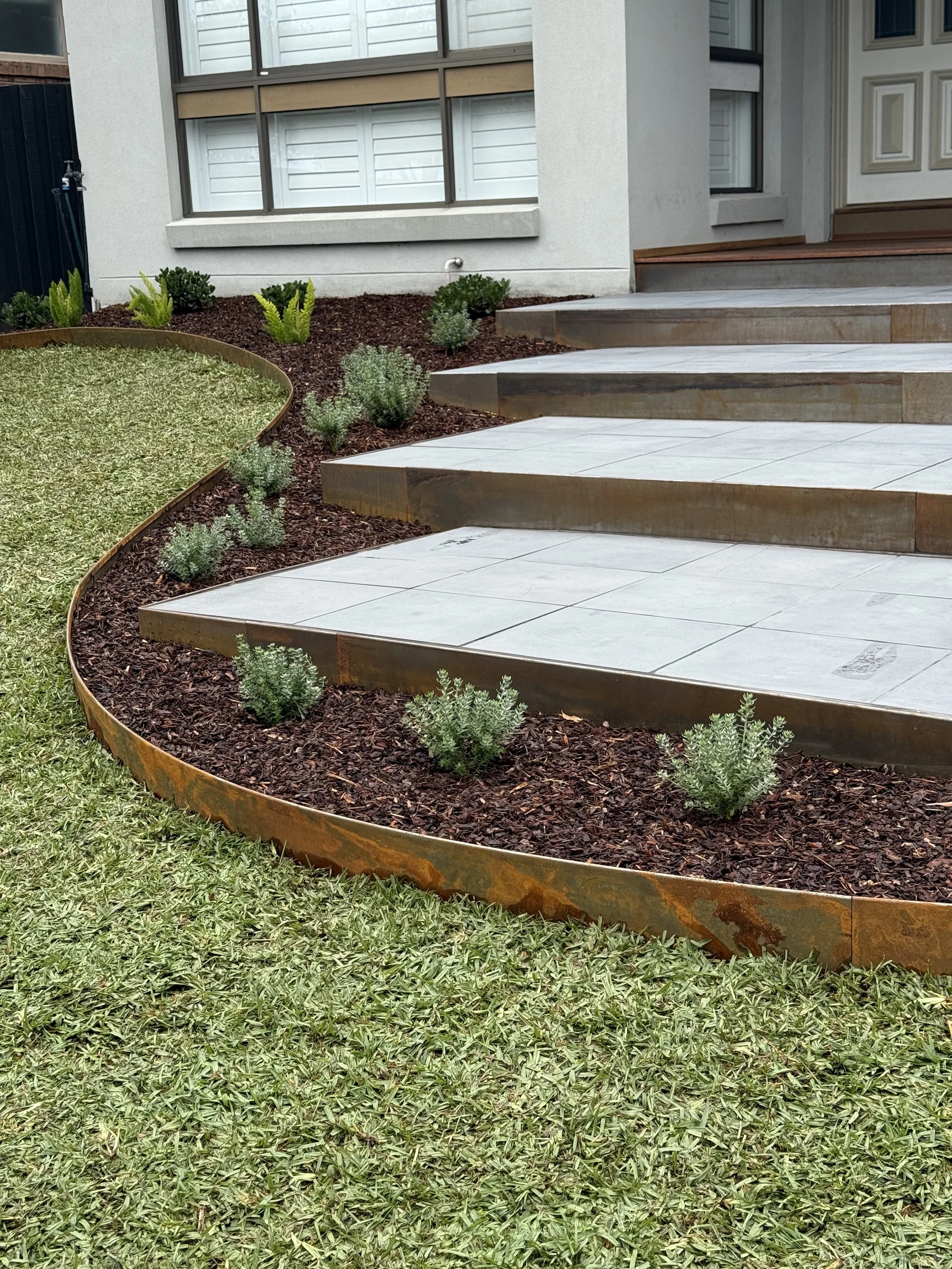 Freshly landscaped front yard with a curved flower bed bordered by rusted metal, containing small plants and mulch, with a concrete step leading to the house entrance and a lawn in the foreground.