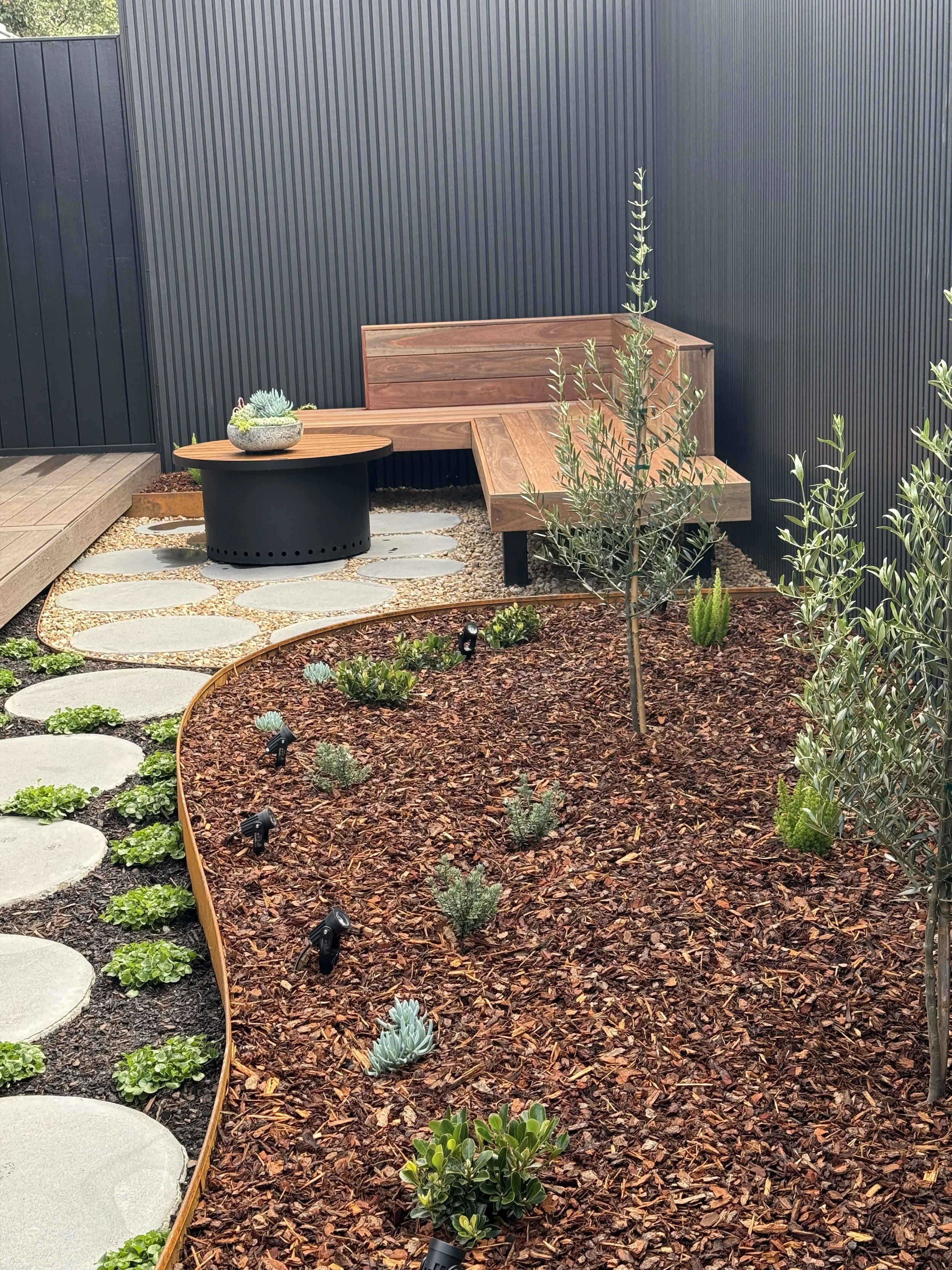 A landscaped backyard garden with a stepping stone pathway, small plants, and a wooden seating area against a black vertical panel fence.