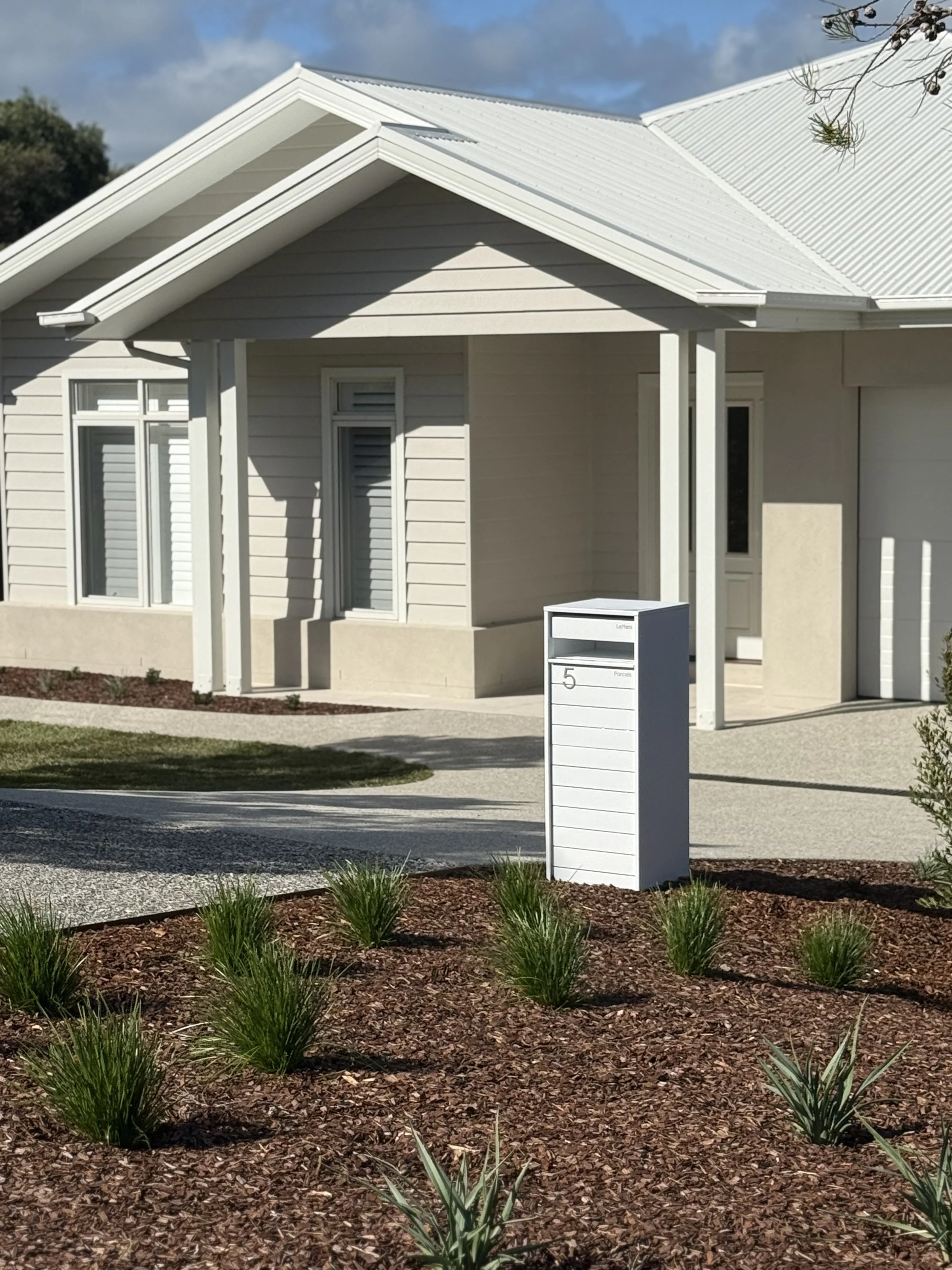 A white residential house with a metal roof, a mailbox labeled the number 5, and a landscaped front yard with small plants and mulch.