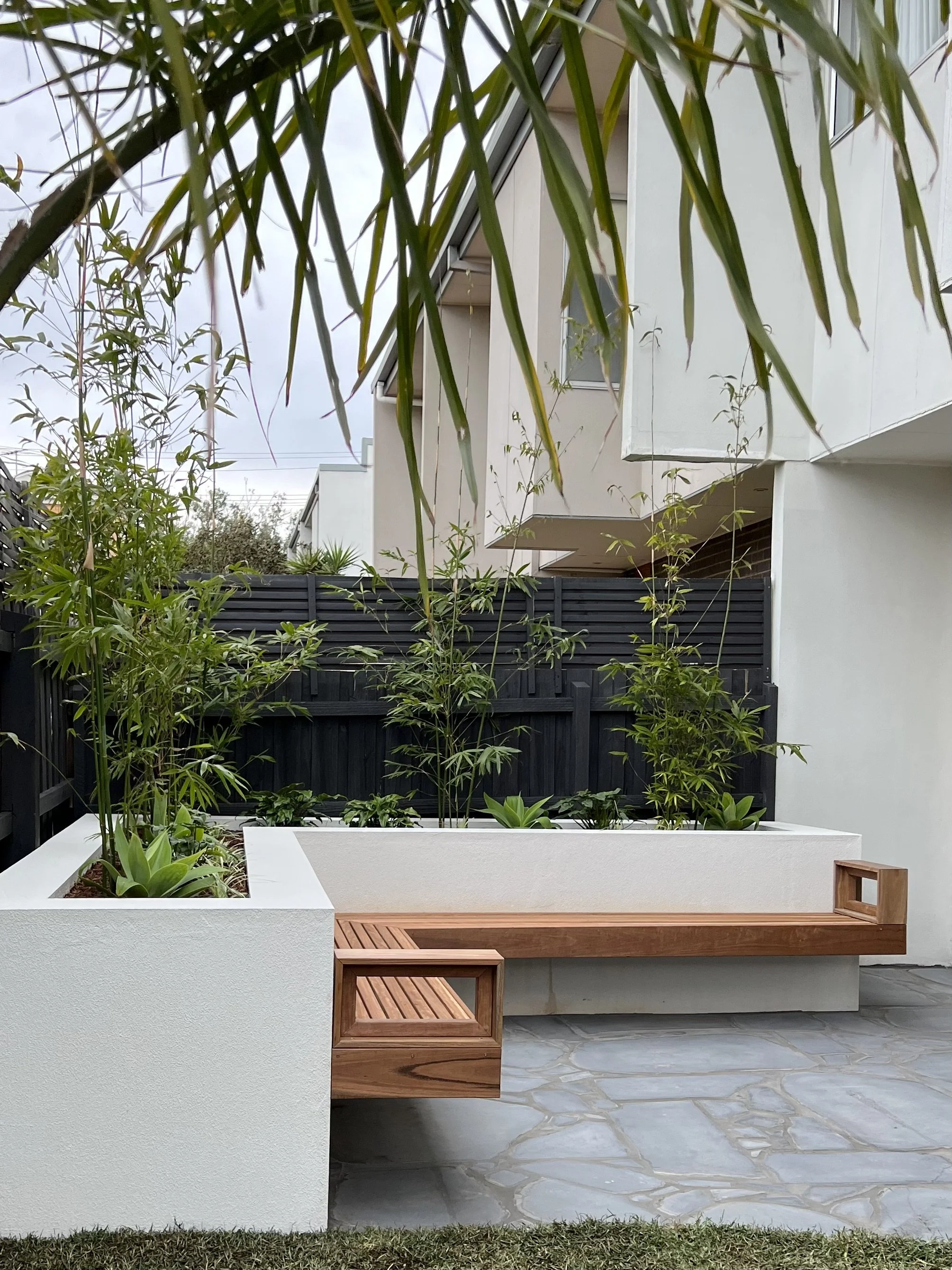 Modern house exterior with white planters, green plants, wooden accents, black fence, gray tiled patio, and a multi-story building in the background under cloudy sky.