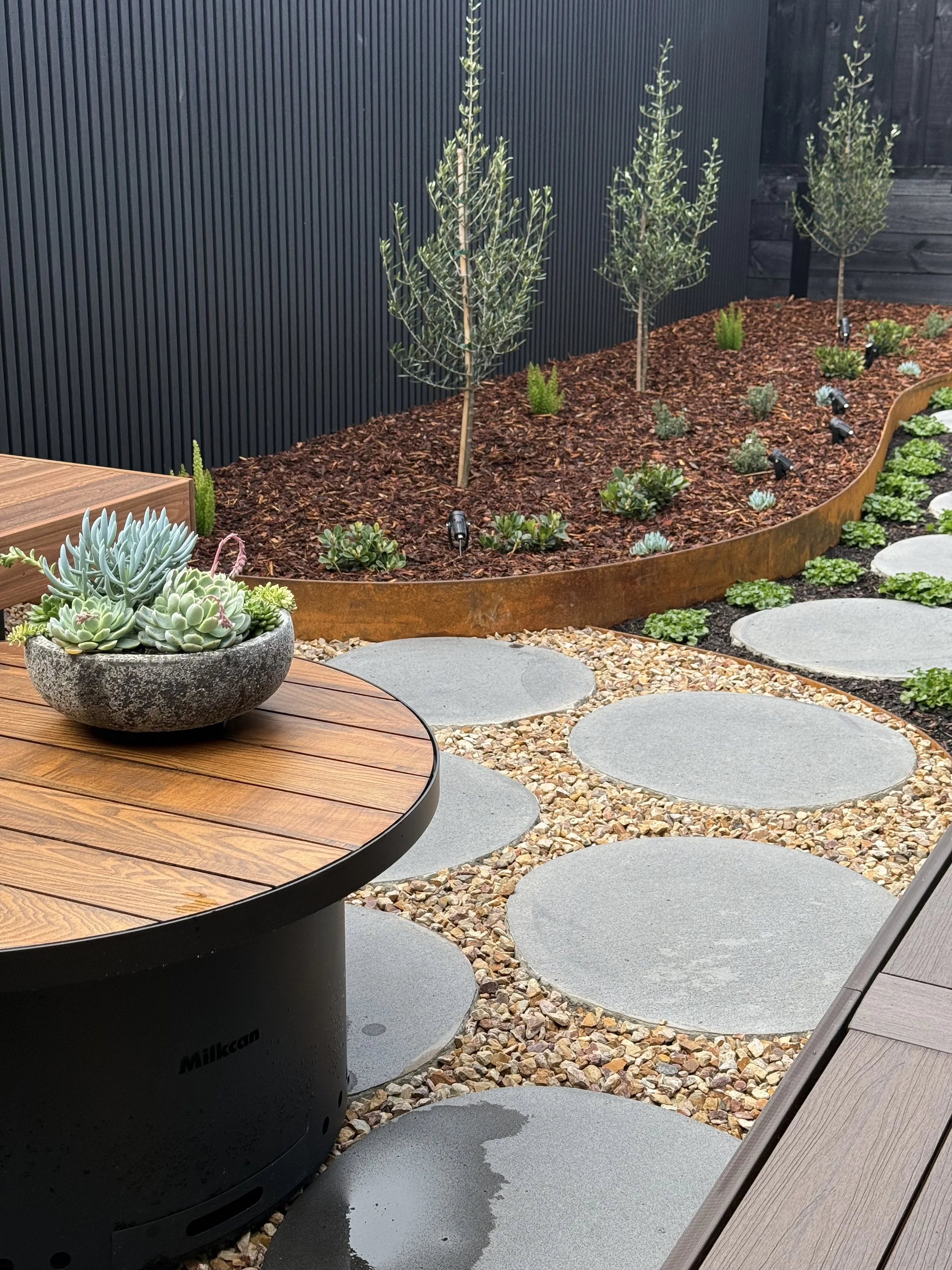 A modern outdoor garden patio featuring a wooden table with a round black base, potted succulents, a gravel pathway with large round stepping stones, a raised garden bed with small trees and plants, a black wall with vertical slats, and a dark fence.