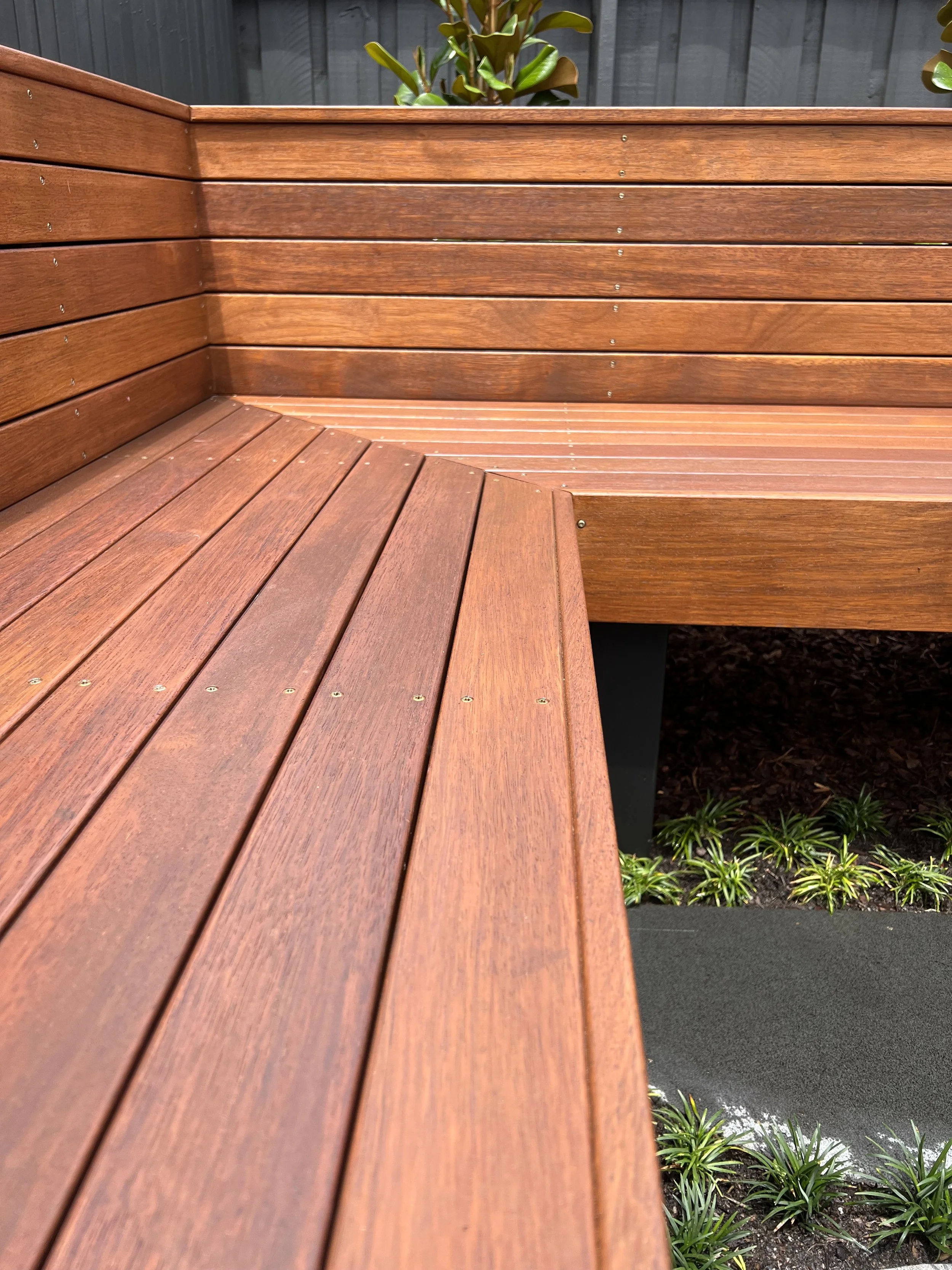 Wooden outdoor seating area with a corner, nearby plants, and a black fence in the background.