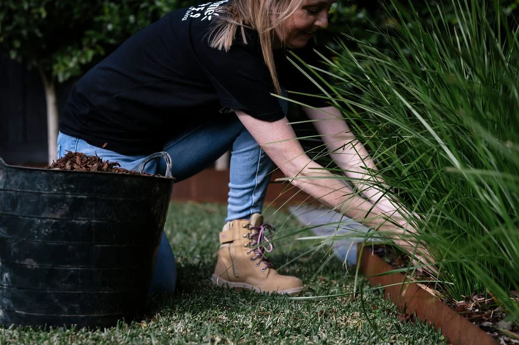 A woman gardening, planting bulbs or plants in a flower bed, with a black container of mulch nearby.