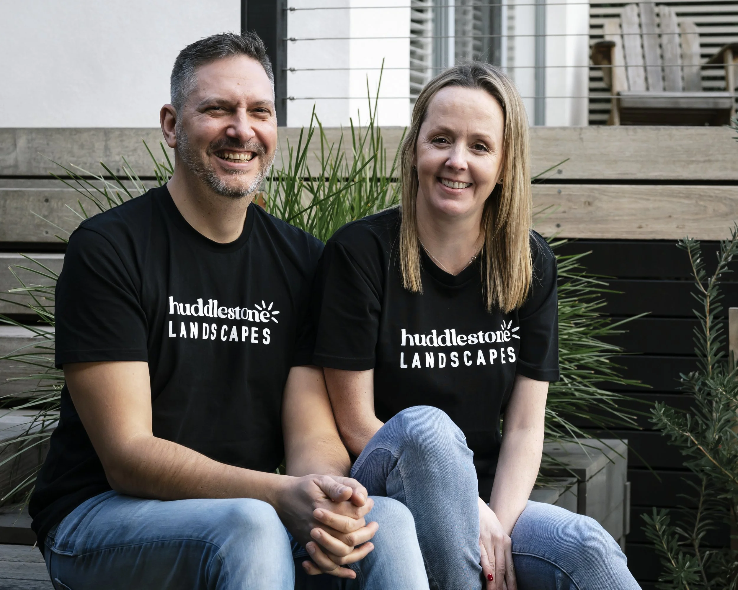 A smiling man and woman sitting together outdoors in front of a wooden fence and green plants. They are wearing black shirts with white text.