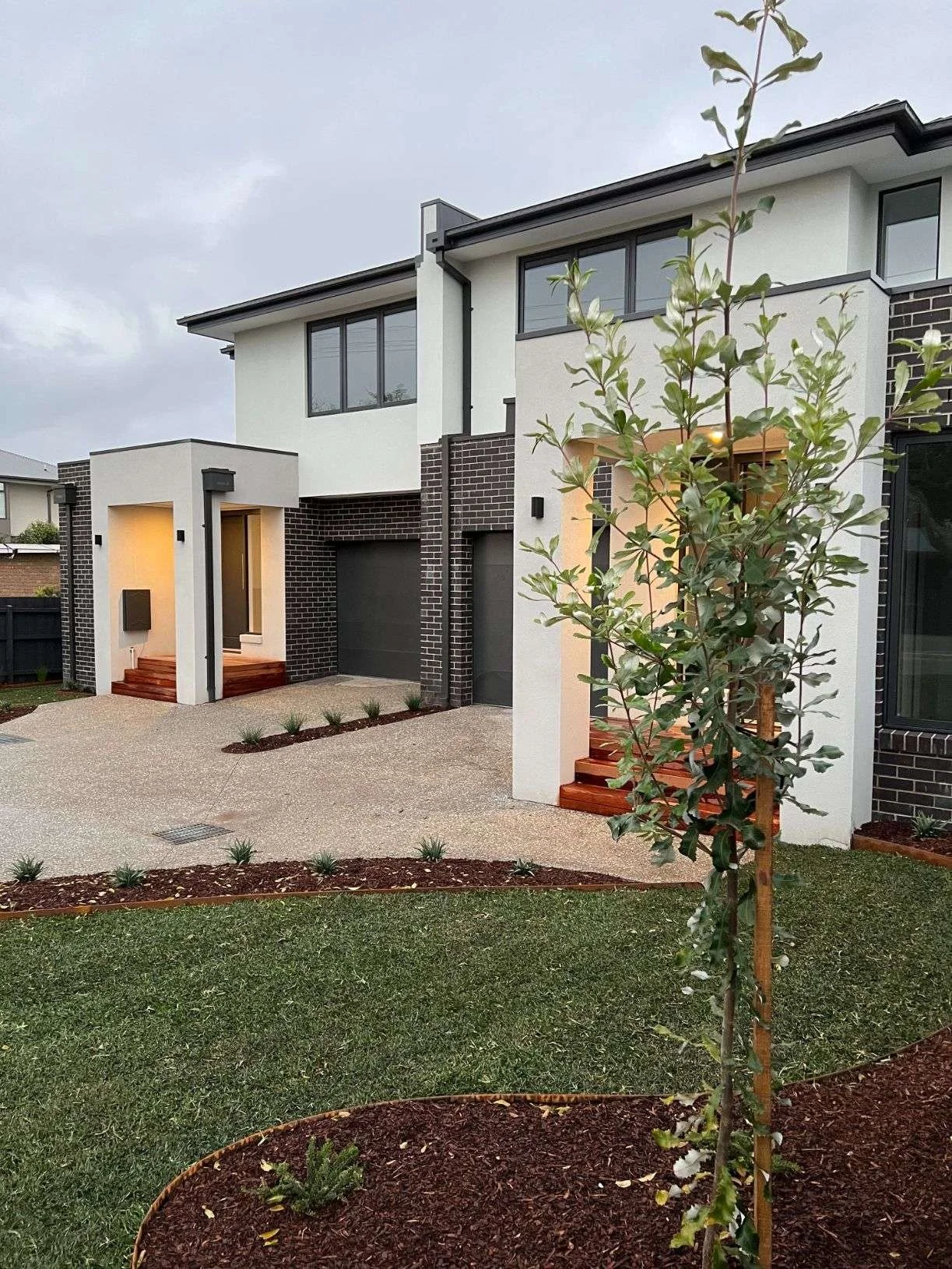 Modern two-story house with white and black brick exterior, small front yard with young tree, gravel driveway, and landscaping with small plants.