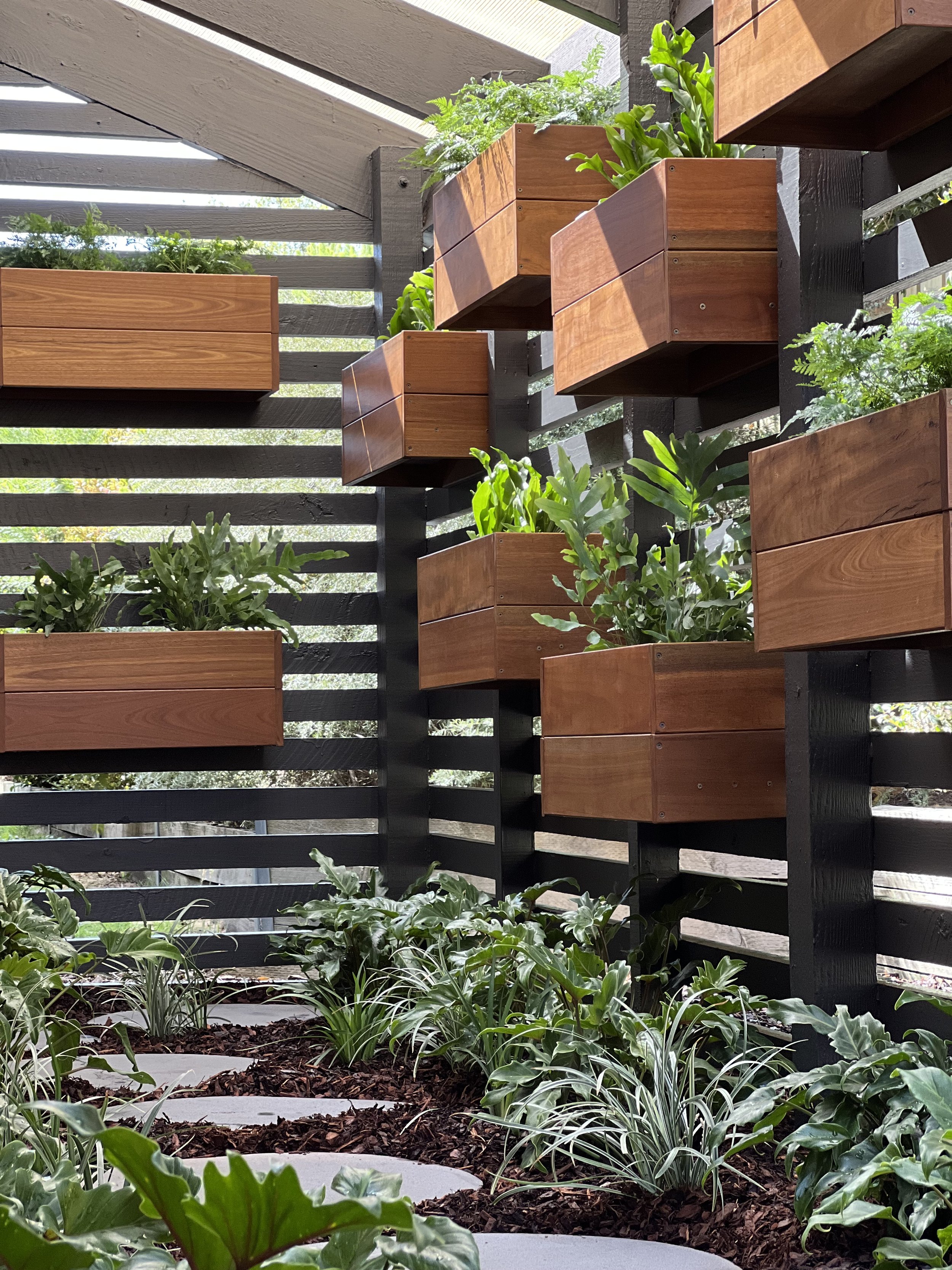A garden with green plants in rectangular wooden planters attached to a black slat fence, with some plants on the ground and sunlight shining on the planters.