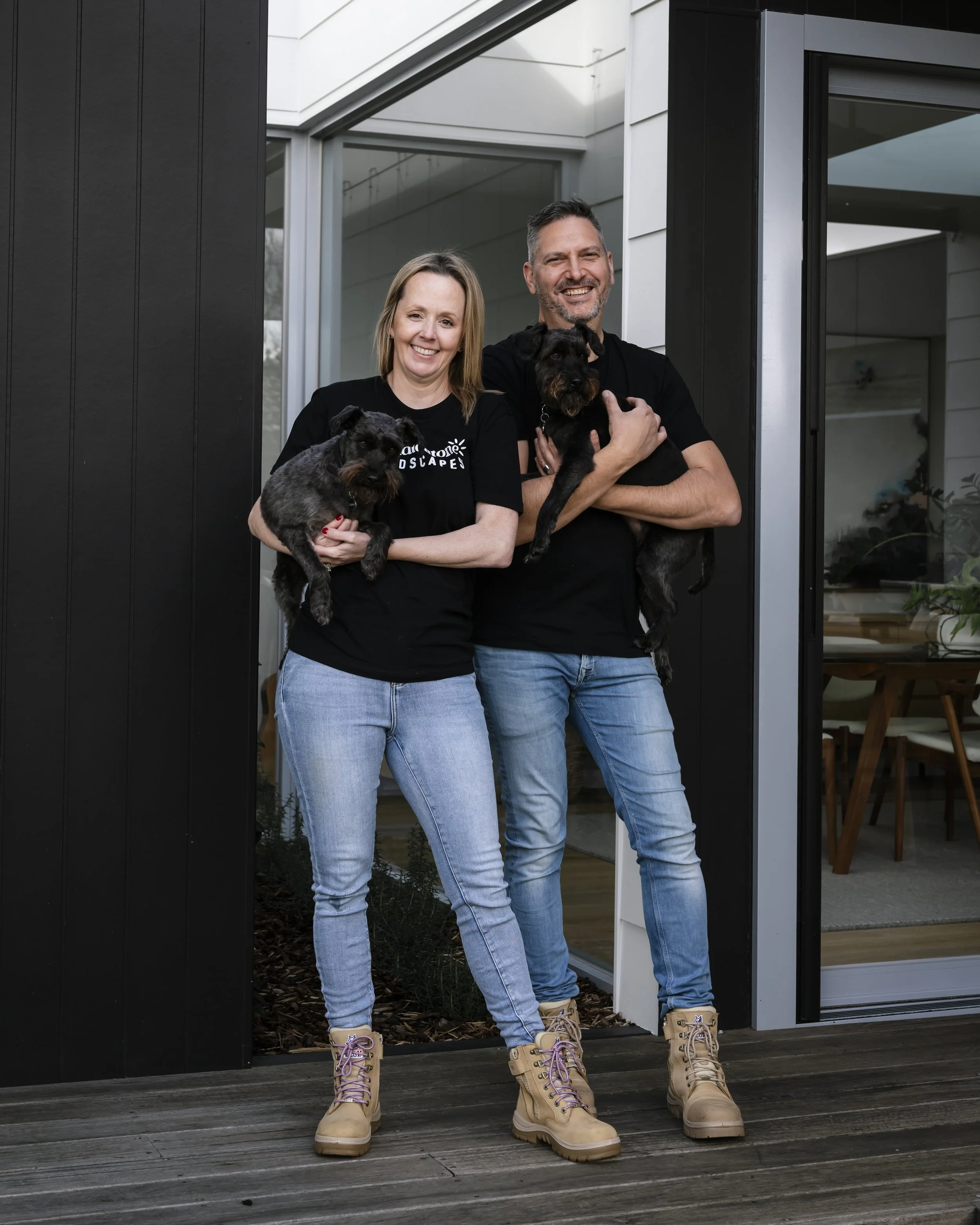 A smiling man and woman holding dogs stand on a wooden porch outside a modern house.