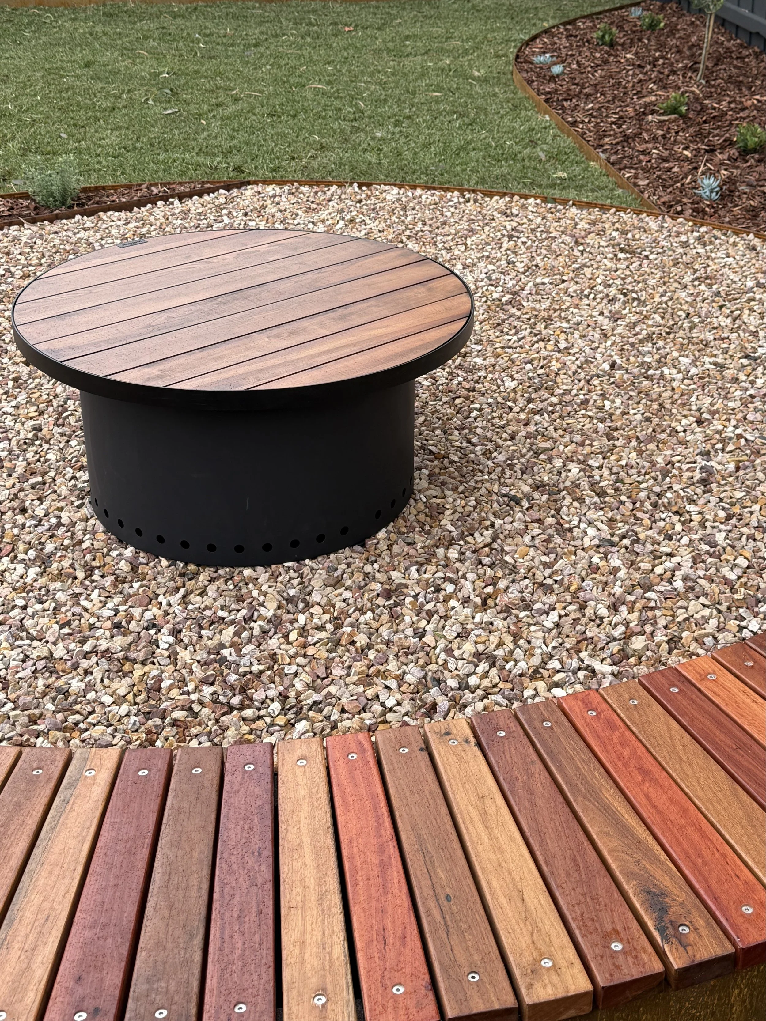 A backyard area with a circular wooden bench on a gravel surface, bordered by a curved flower bed with small succulents and a patch of grass.