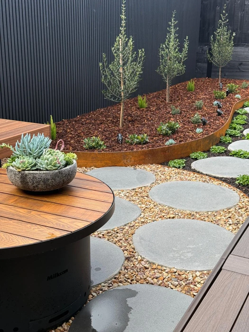 A modern outdoor garden patio featuring a wooden table with a round black base, potted succulents, a gravel pathway with large round stepping stones, a raised garden bed with small trees and plants, a black wall with vertical slats, and a dark fence.