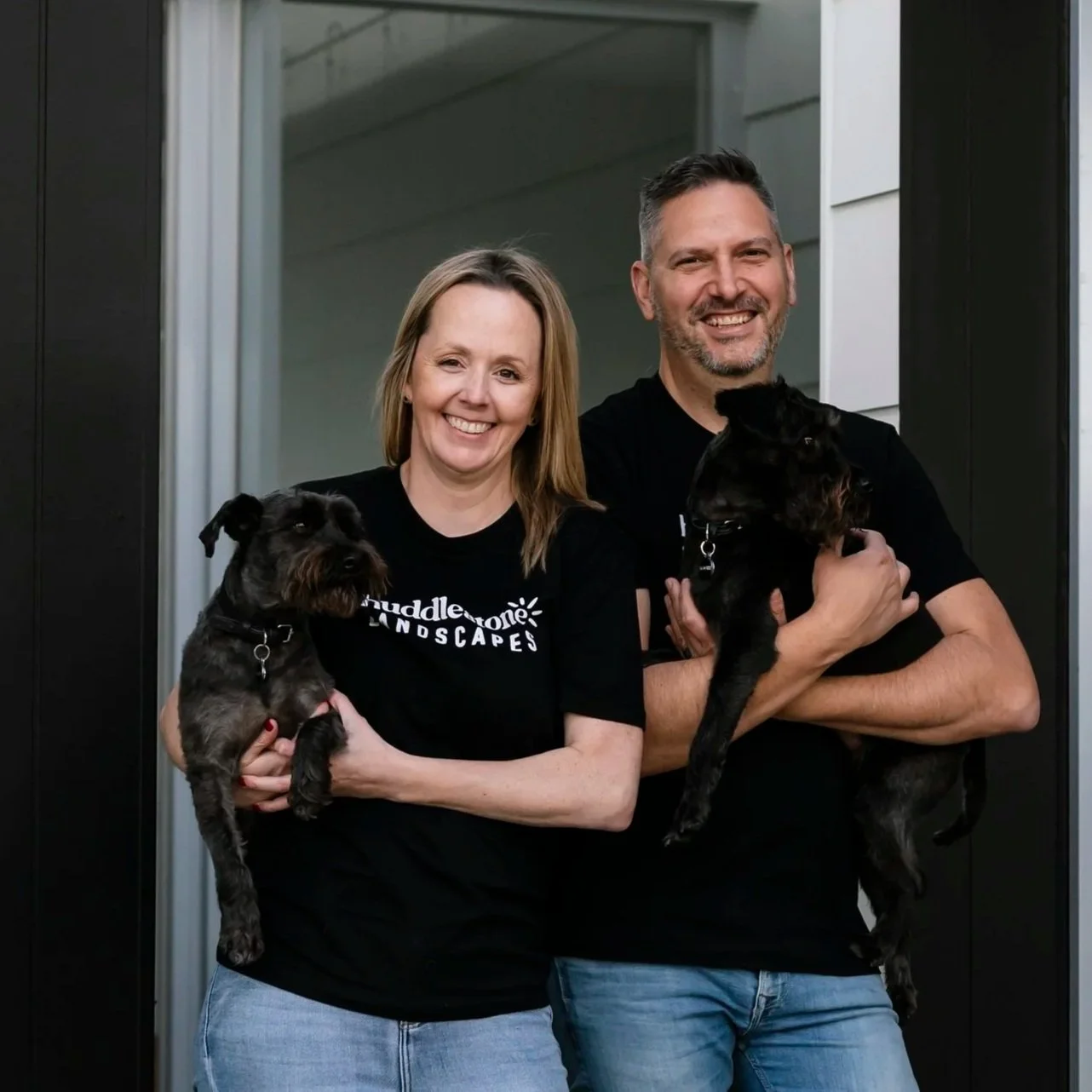 A smiling man and woman standing outside a house, each holding a small black dog. The woman wears a black shirt with white text, and the man wears a plain black shirt. Both are wearing jeans.