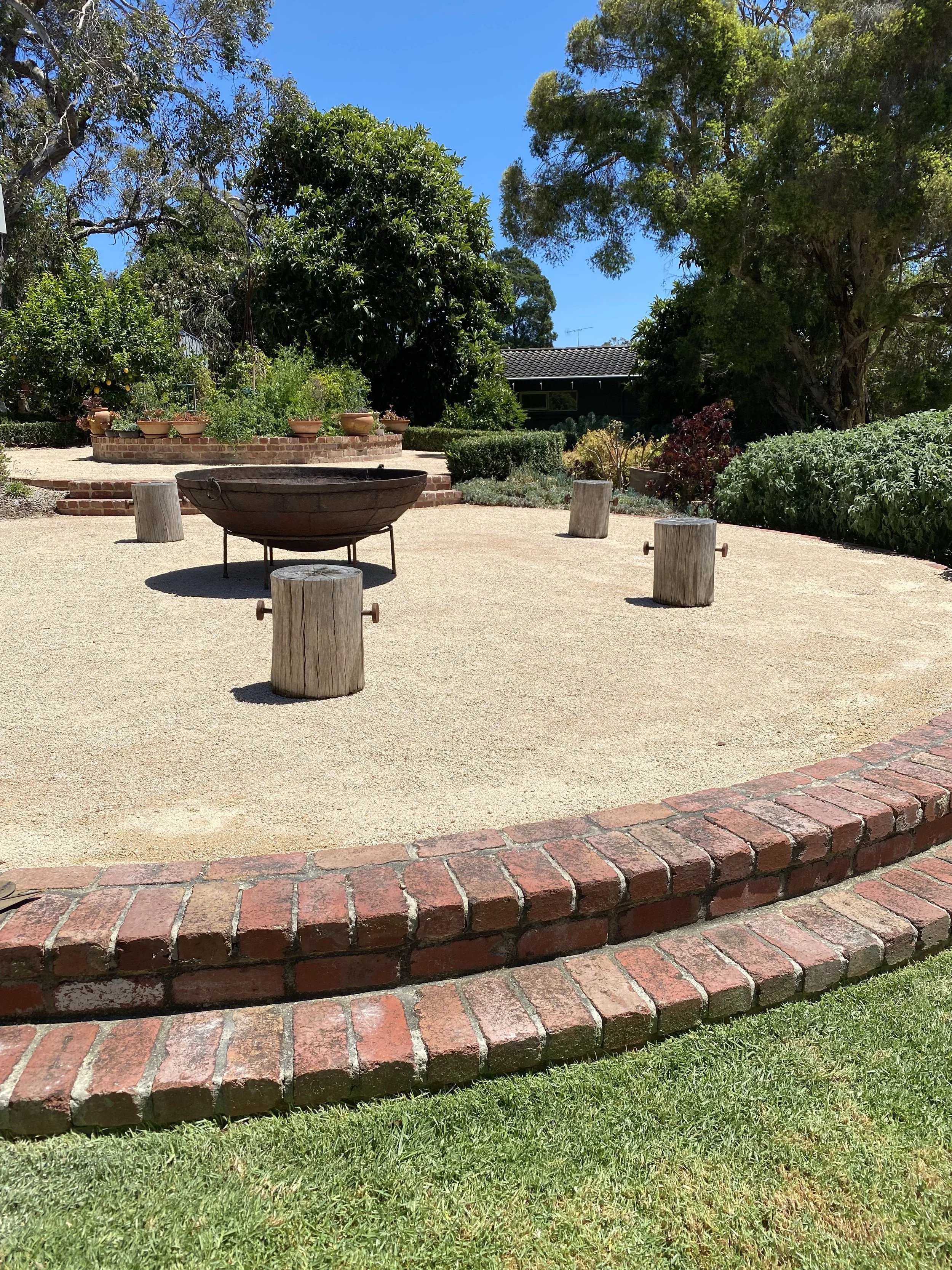 A garden area with a large round metal fire pit on a concrete surface, surrounded by four wooden stumps, and a brick border with a grassy lawn outside. There are trees and bushes in the background under a bright blue sky.