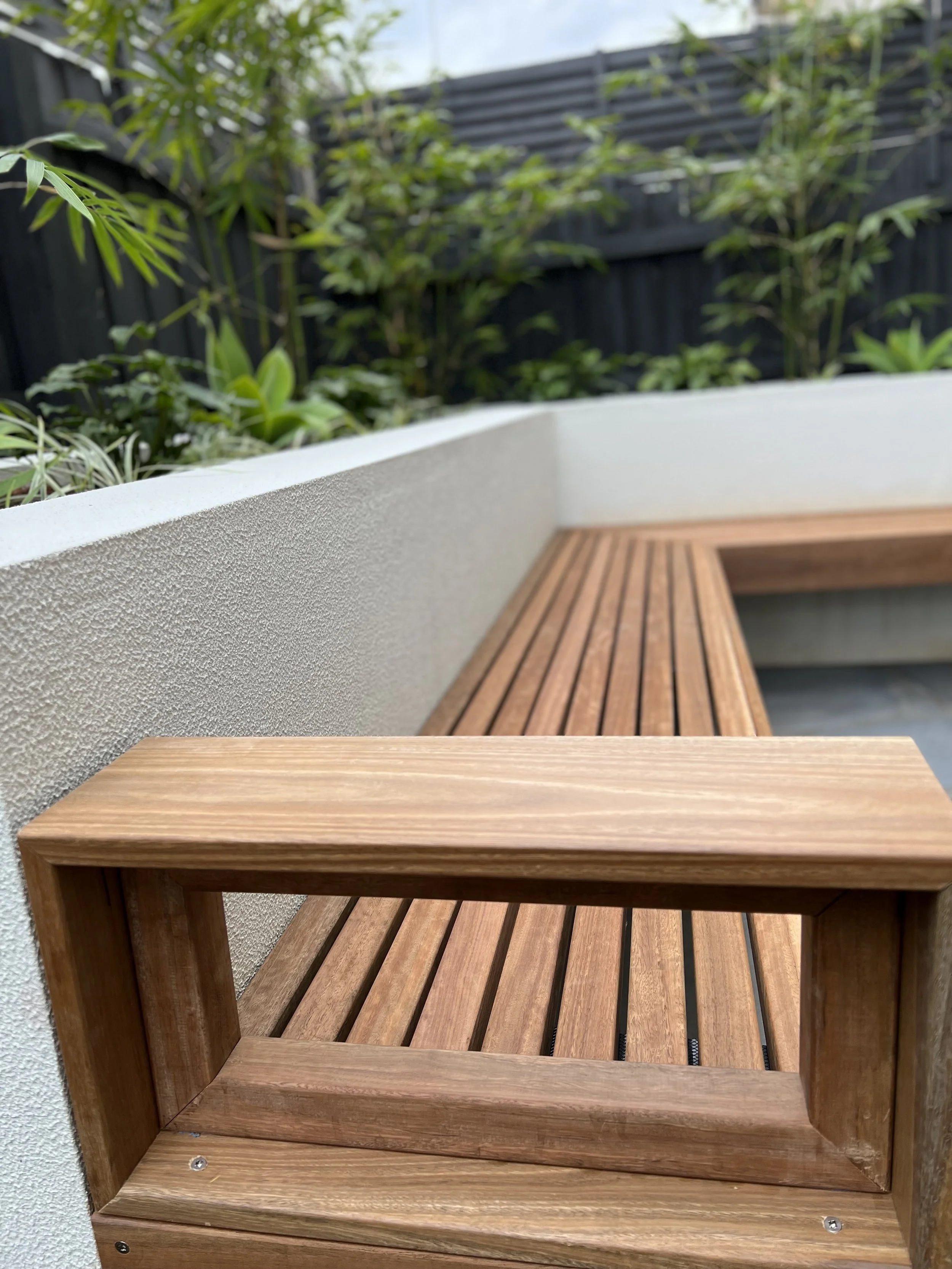 Close-up of a wooden bench on a patio, with a textured white wall and green plants and shrubbery in the background.