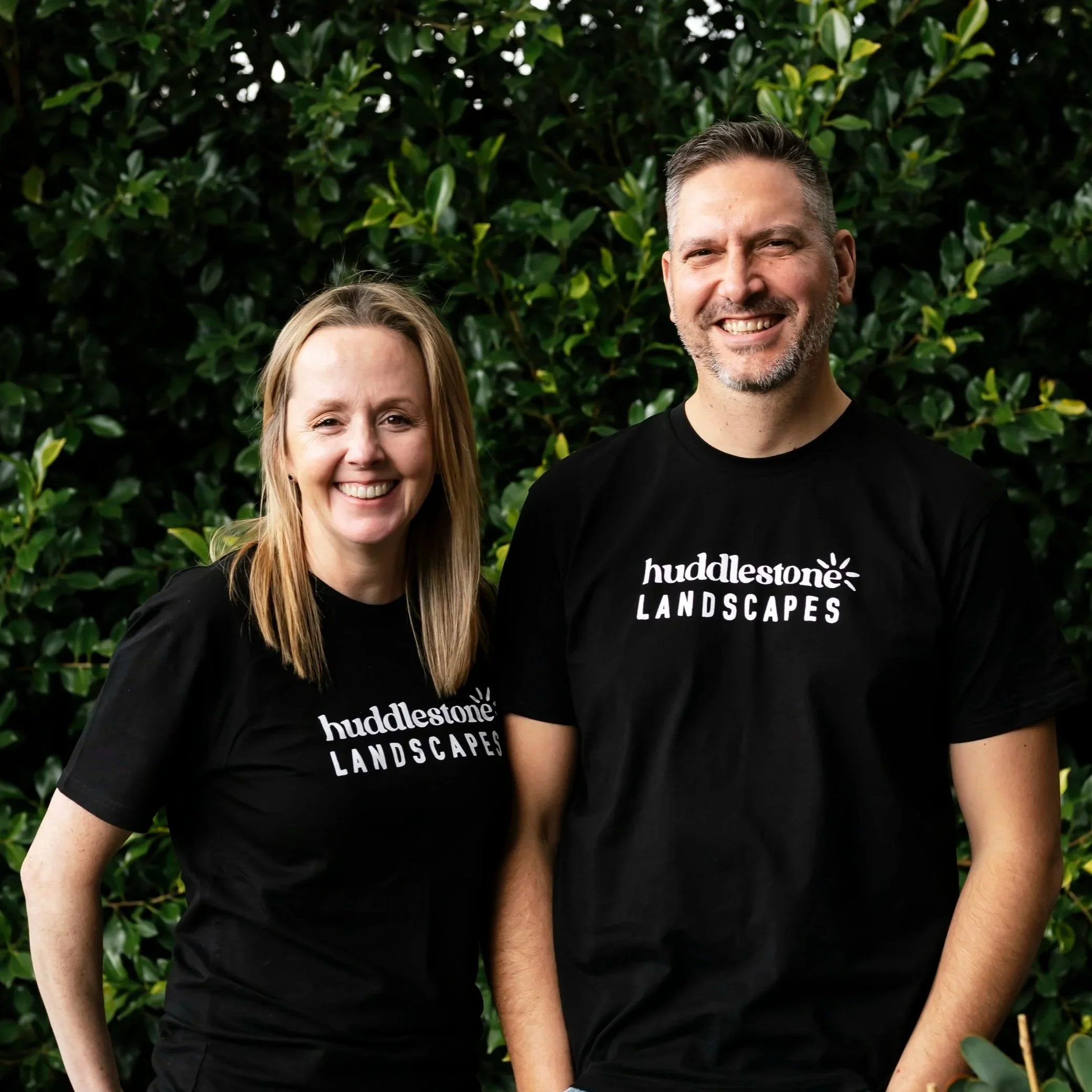 Smiling woman and man wearing black t-shirts with 'Huddletone Landscapes' logo, standing outdoors in front of green leafy bushes.