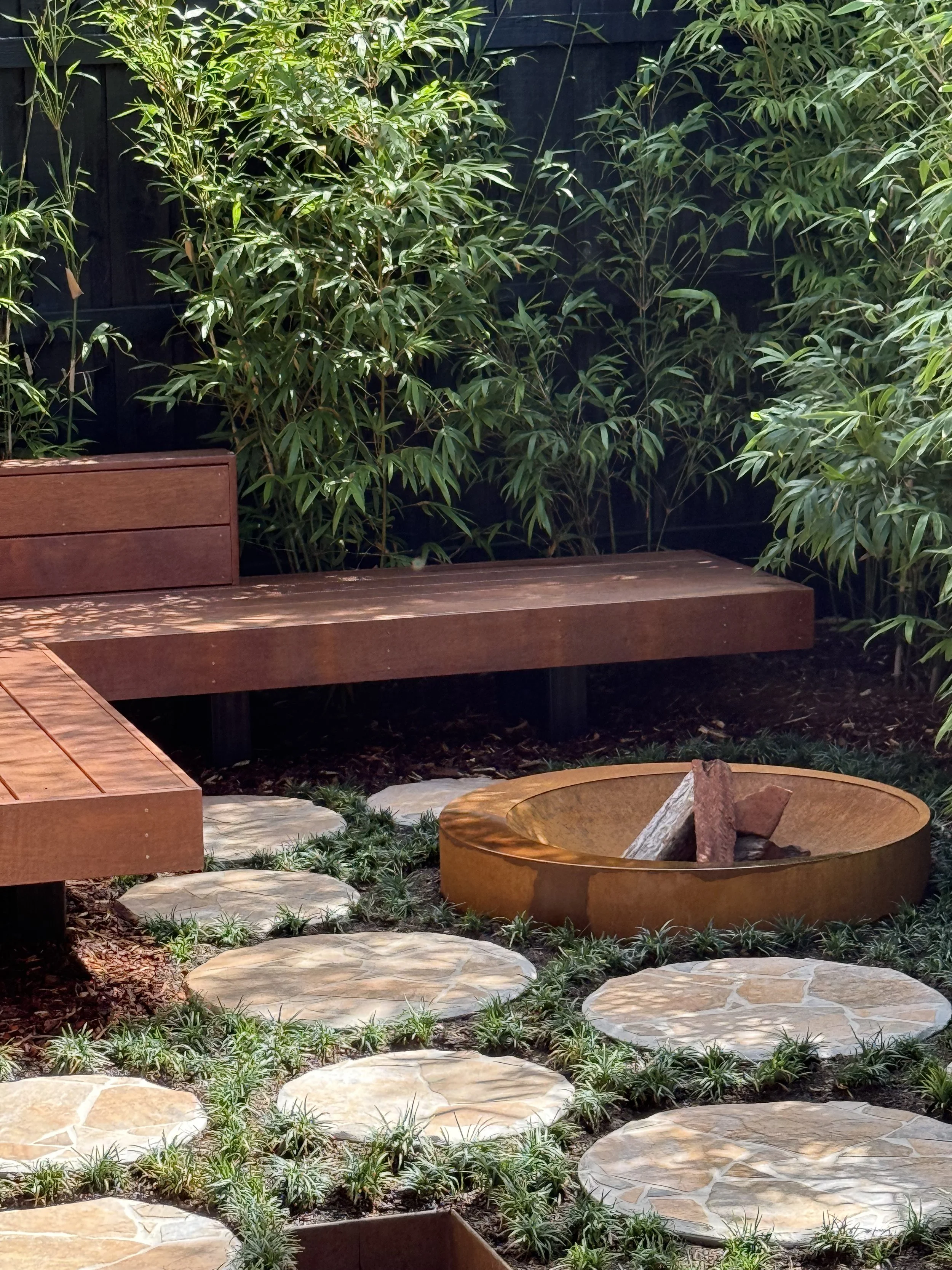 A serene outdoor garden scene with a wooden bench, a circular fire pit with logs inside, stone pathway, lush green foliage, and a dark fence in the background, illuminated by soft lighting.