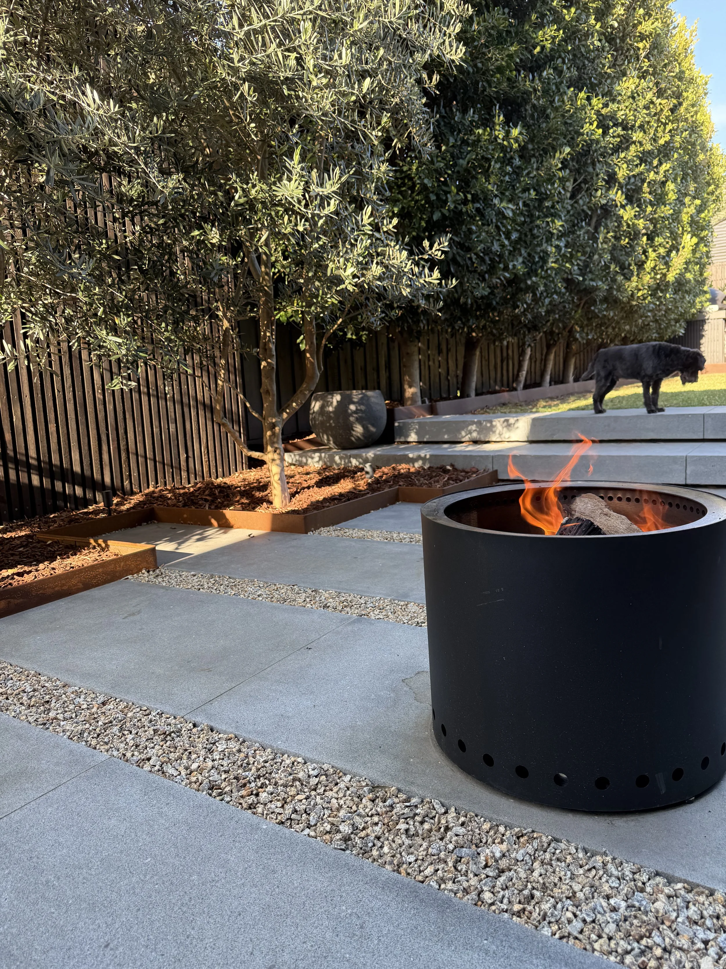 A backyard with a fire pit, concrete and gravel patio, a wooden fence, trees, and a black dog standing on the steps. The scene is lit by sunlight, with a clear blue sky visible.