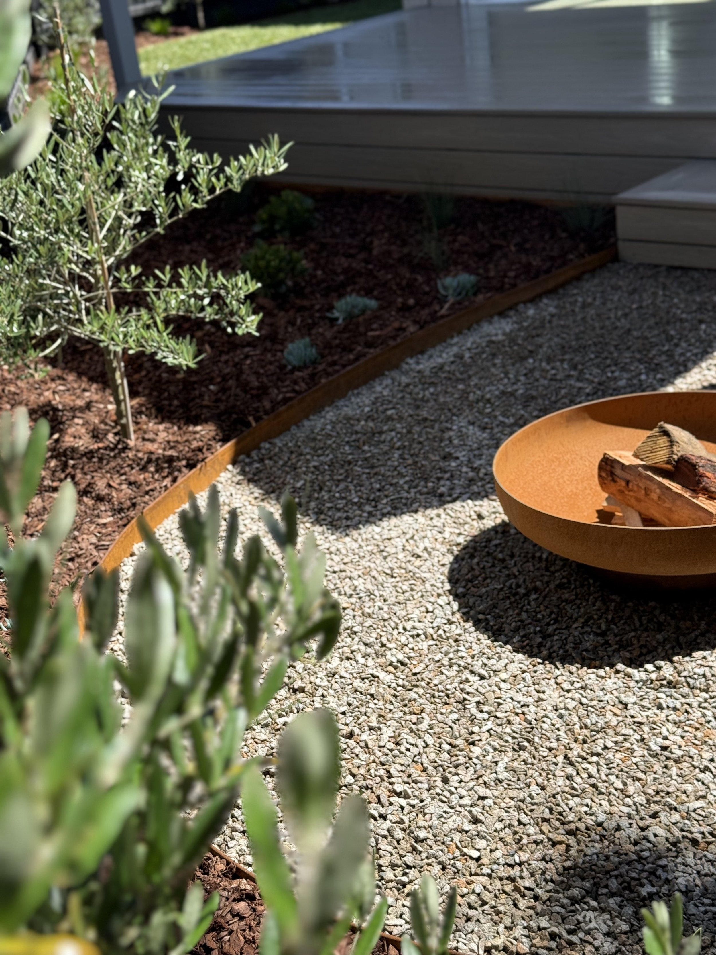 A landscaped backyard with a gravel pathway, a curved wooden border, green plants, and a grey deck. There is a rusty firepit in the foreground.