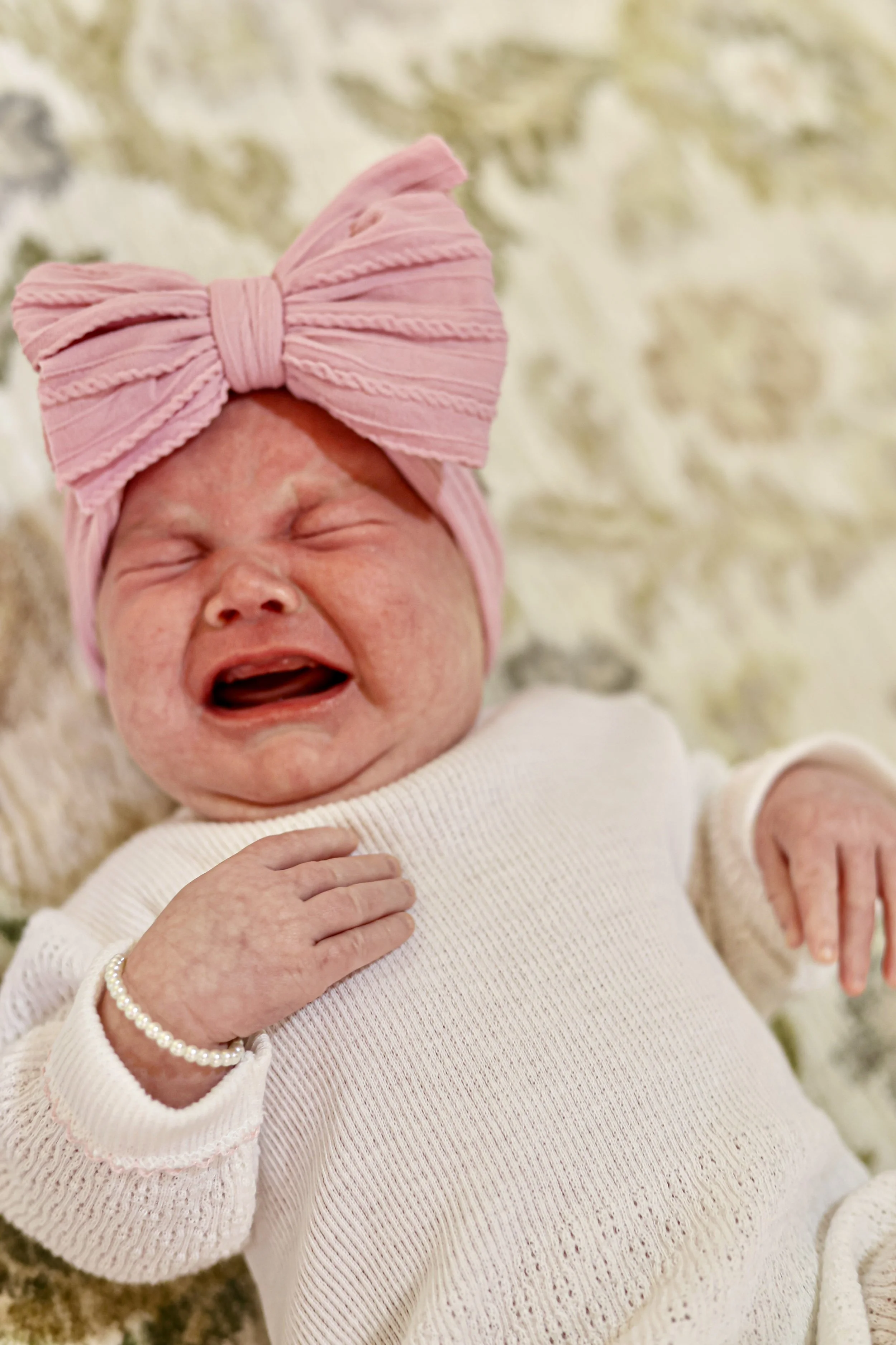 Close-up of a crying baby girl with a pink headband, wearing a white sweater, holding her chest with her hand, on a soft beige surface.
