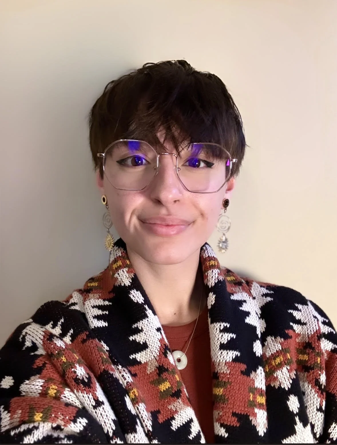 A person with short dark hair, glasses, and earrings, smiling, wearing a colorful patterned jacket and a necklace, standing against a plain light-colored wall.