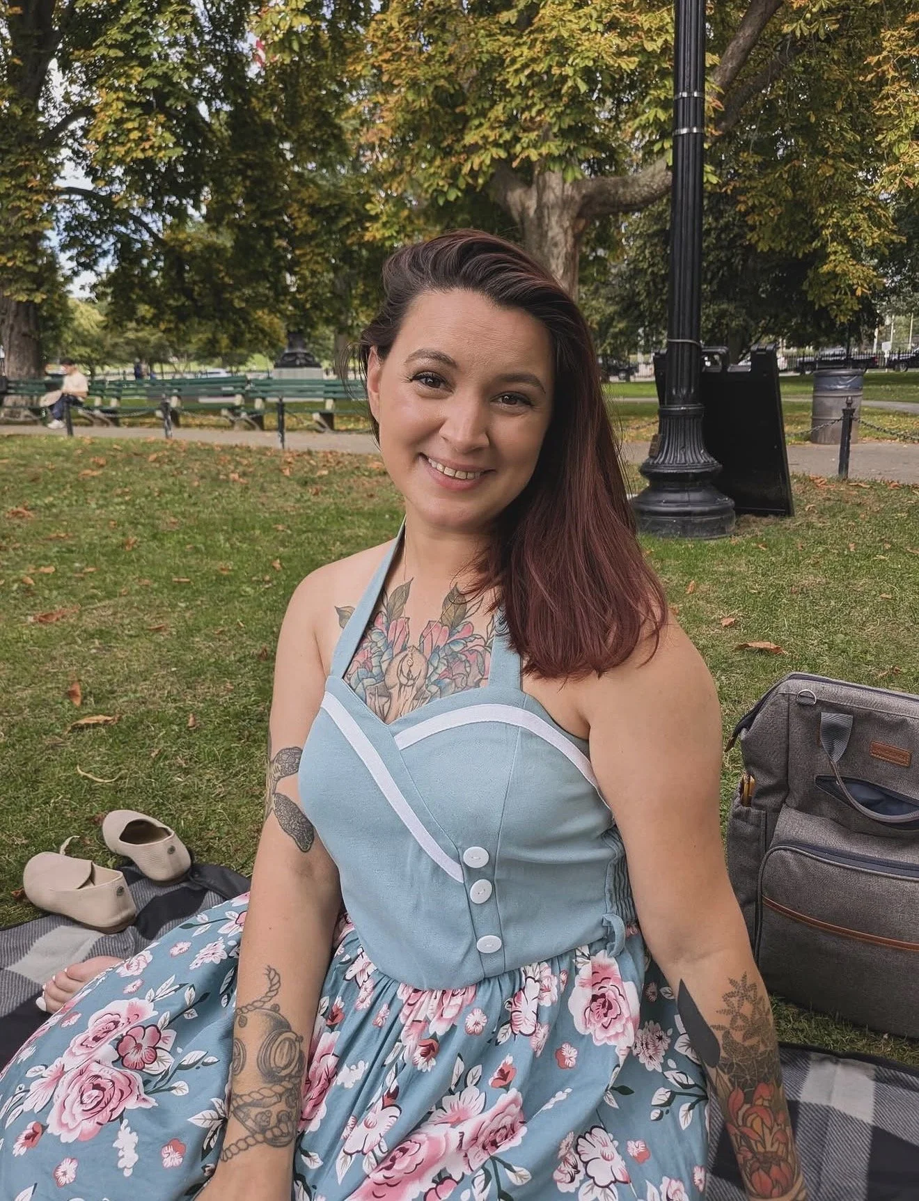 A smiling woman with tattoos on her arms and chest sitting on a picnic blanket in a park, wearing a floral dress with a light blue top. There are shoes and a backpack on the blanket. Trees and park benches are in the background.