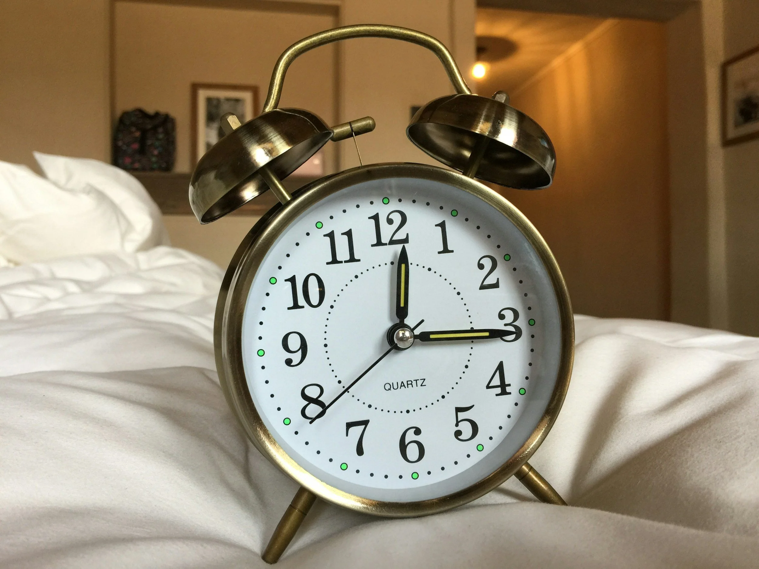 A metal vintage alarm clock showing the time as 3:00, placed on a bed with white sheets, against a background of a room with warm lighting and framed pictures on the wall.