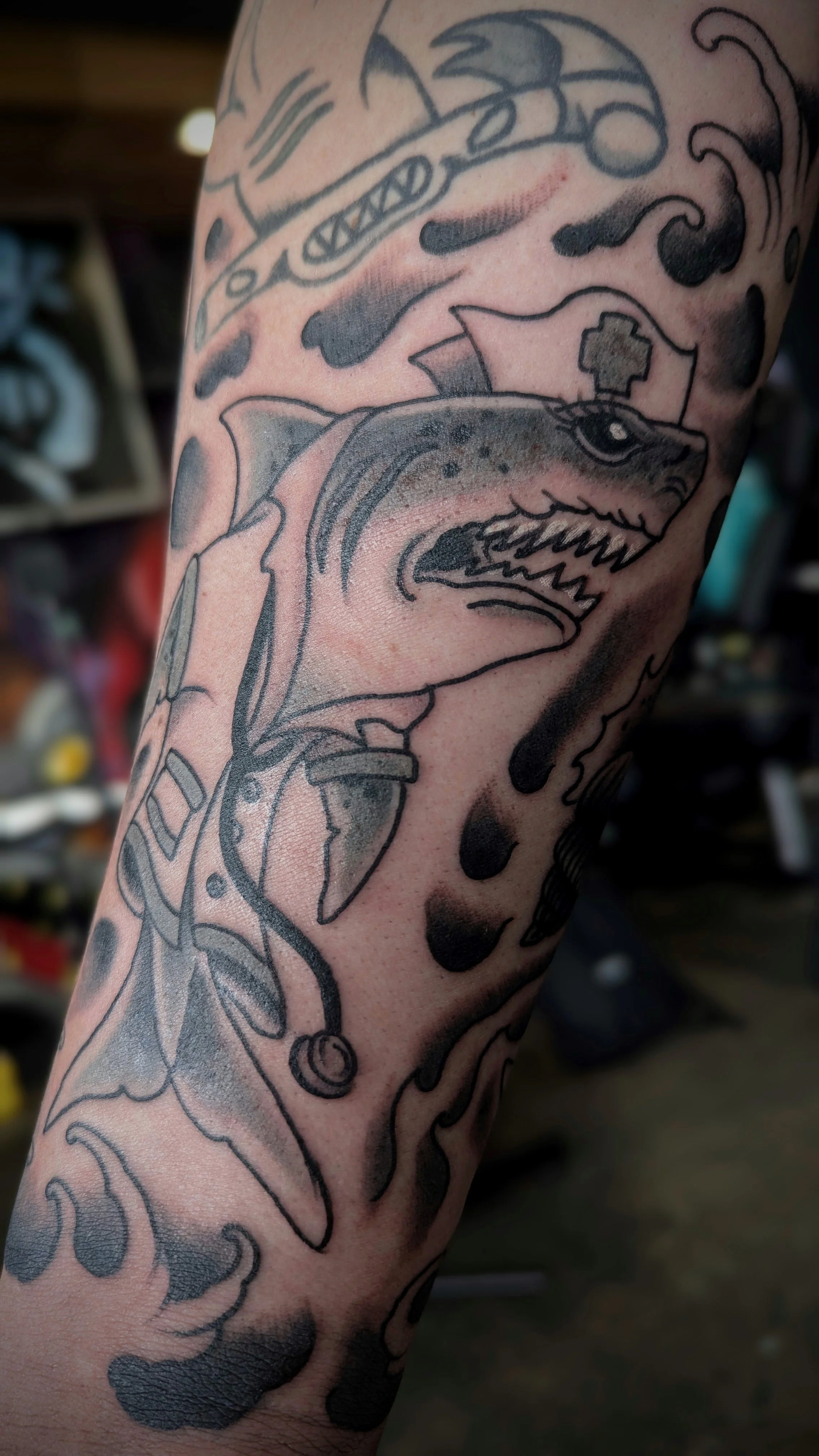 Tattoo of a fish with an open mouth and sharp teeth on a person's arm, surrounded by abstract wave and cloud patterns.