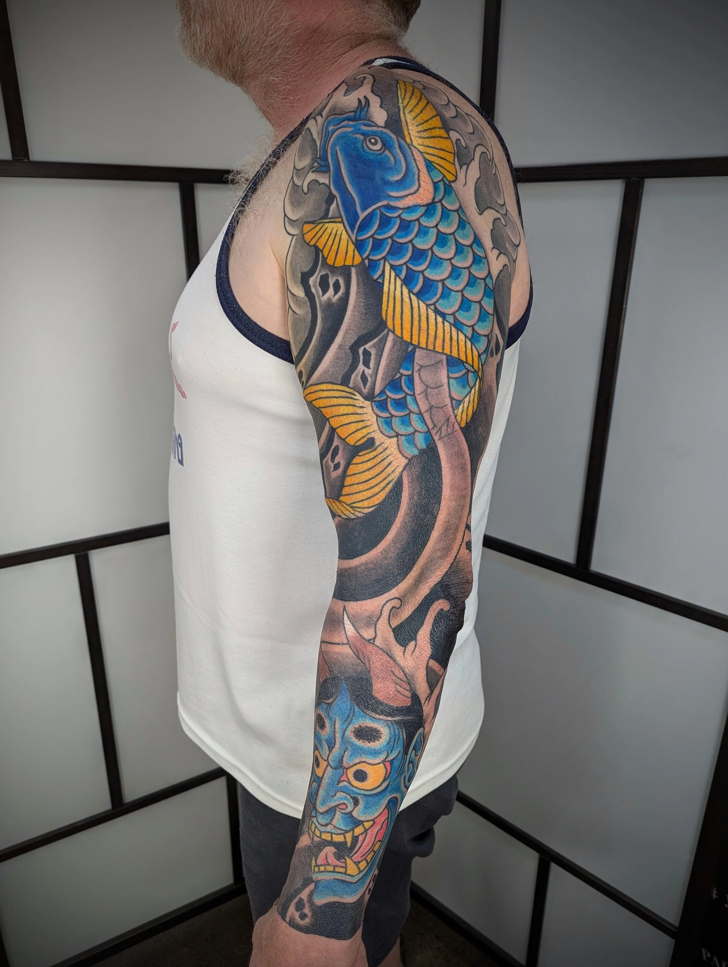 A tattooed man wearing a white sleeveless shirt with dark trim, standing in front of a plain background with black framing.